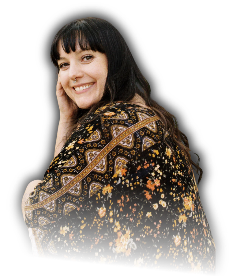 A woman with long dark hair and a nose piercing, smiling and looking over her shoulder, wearing a black floral dress with yellow, orange, and white flowers, against a black background.