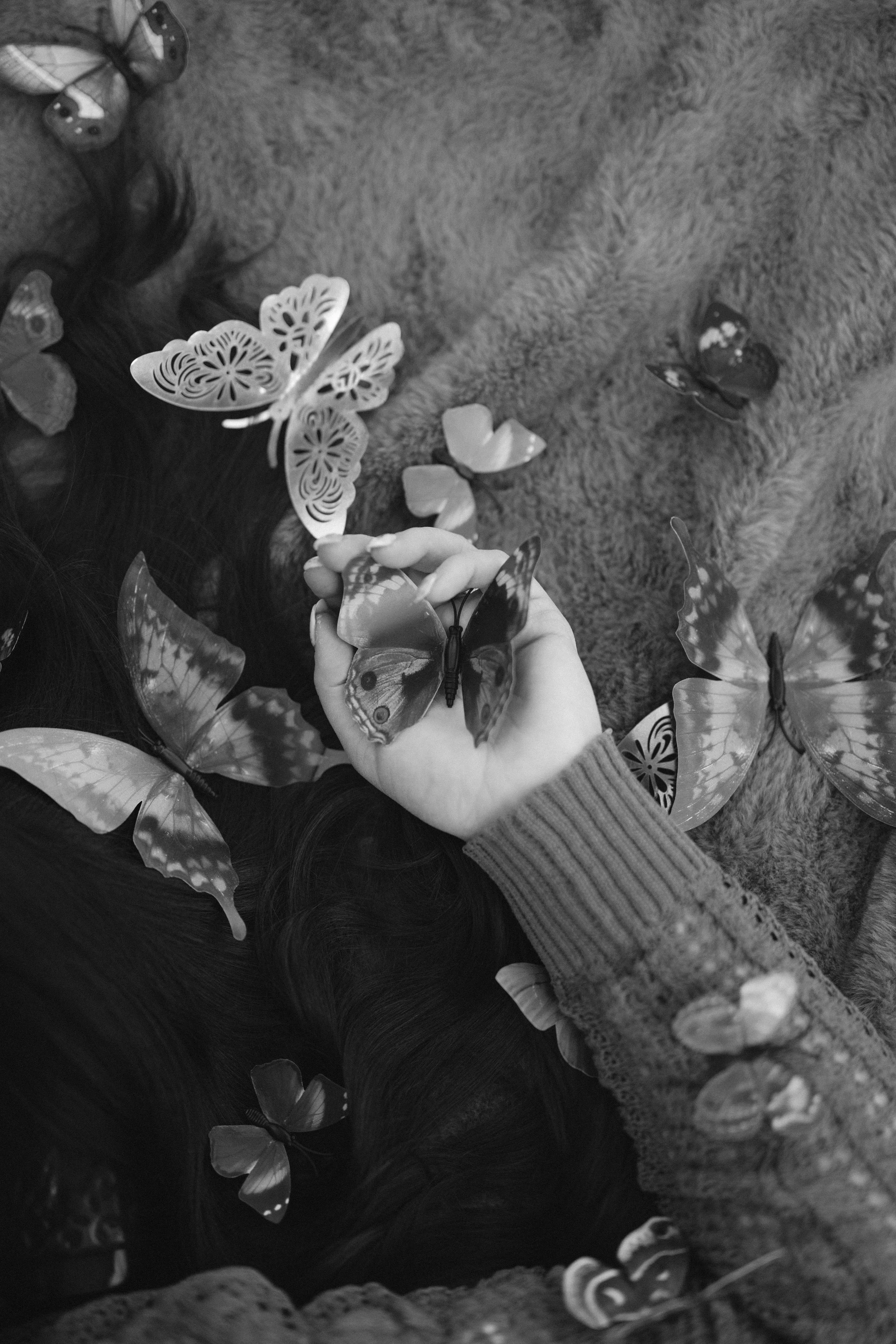 A person with long hair, wearing a sweater, lying on a furry surface, playing with and looking at several artificial butterflies scattered around.