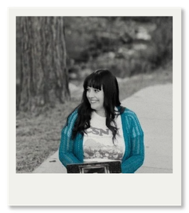 A young woman with long dark hair, wearing a blue jacket, sitting on a park bench, smiling, with trees and a path in the background.