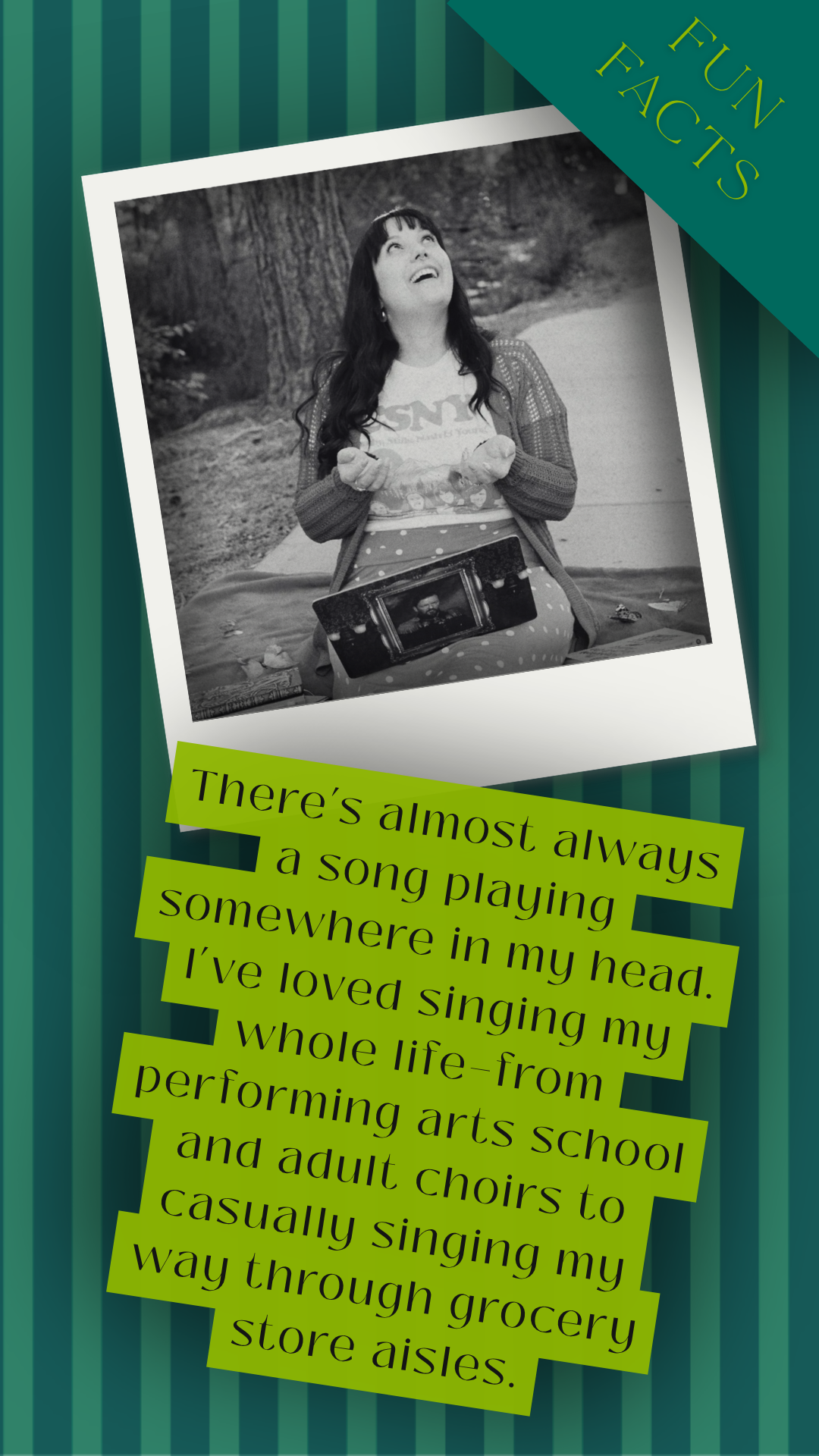 A woman with long dark hair sitting outdoors holding a small object and looking up with a joyful expression. There is a framed photo on her lap and a blanket beneath her. The background shows trees and a pathway.