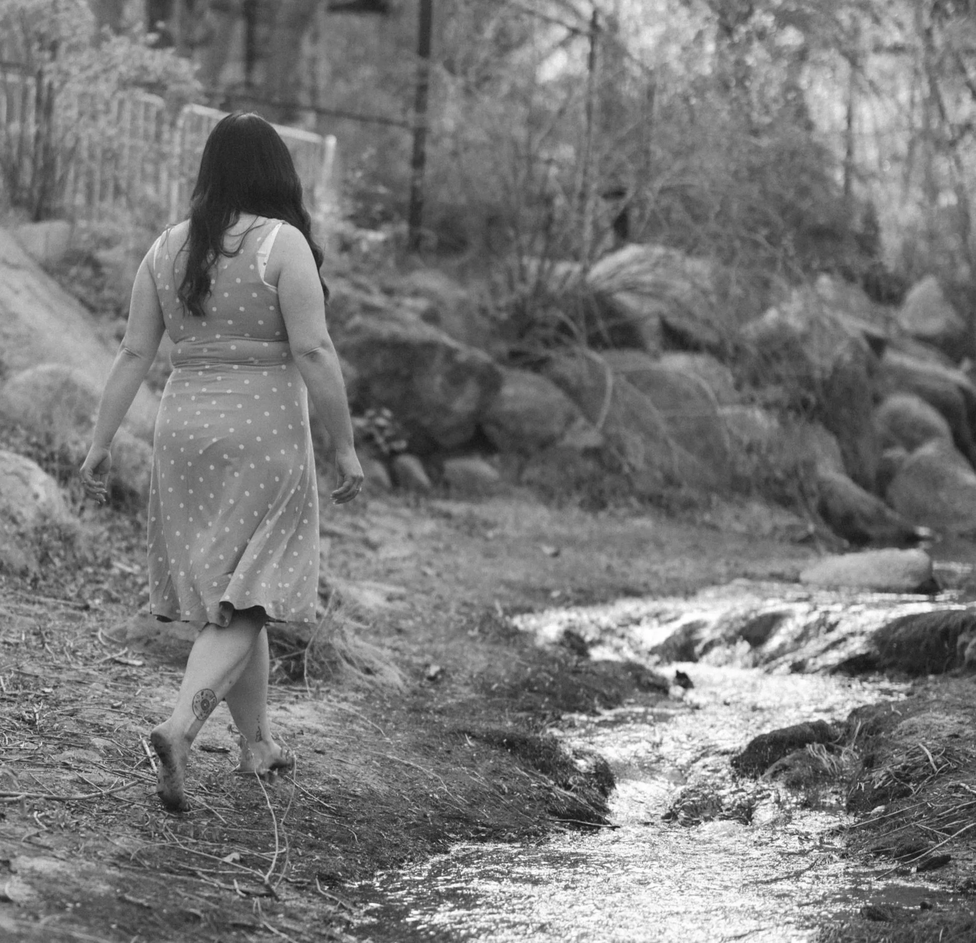 A woman with long dark hair, wearing a polka dot dress, walking barefoot in a natural outdoor setting with rocks, trees, and a stream visible in the background.