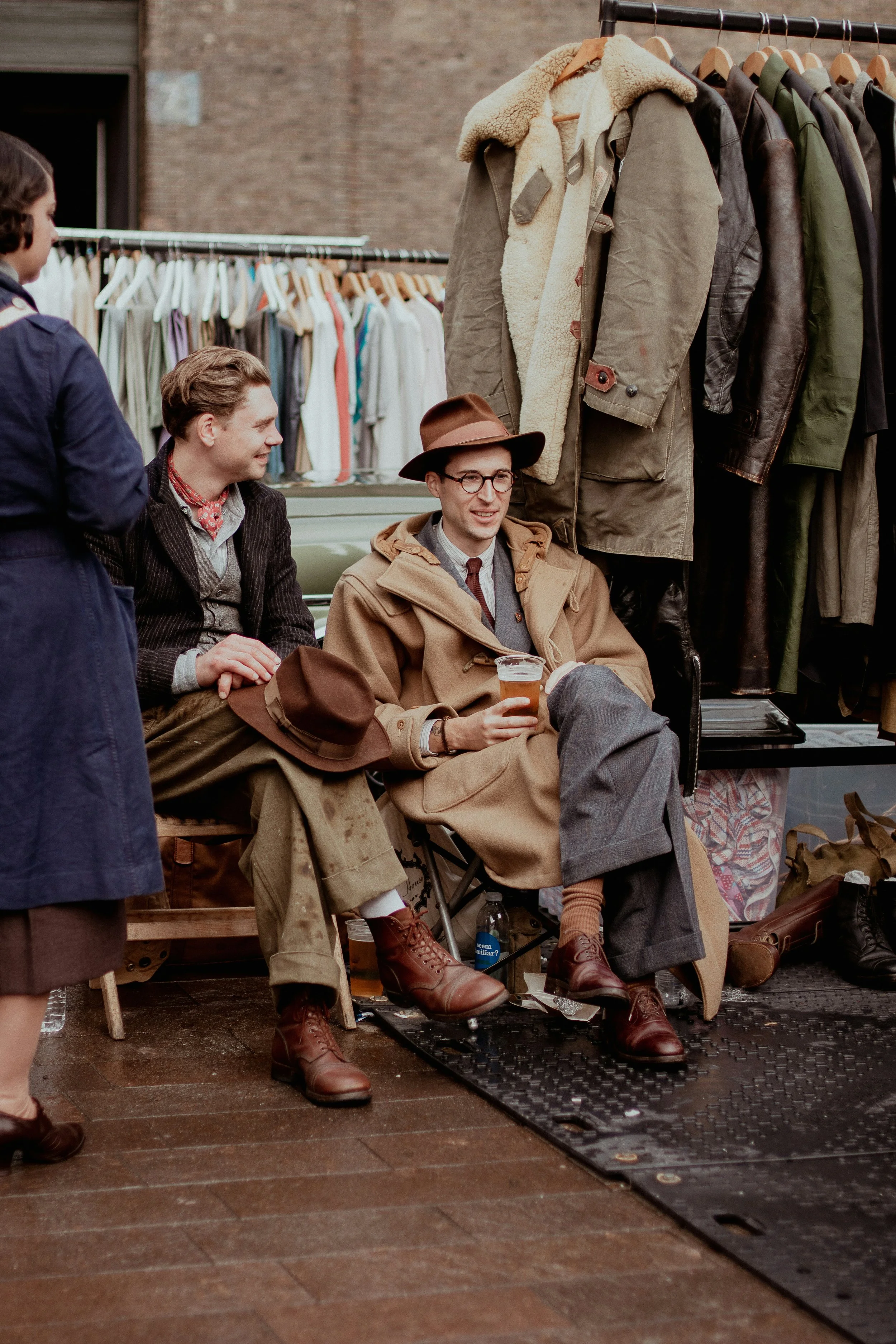 Three men dressed in vintage clothing sitting at a sidewalk market stall with hanging jackets and clothes behind them. They are smiling and conversing, with one man holding a glass of beer. A woman in a blue coat is partially visible on the left.