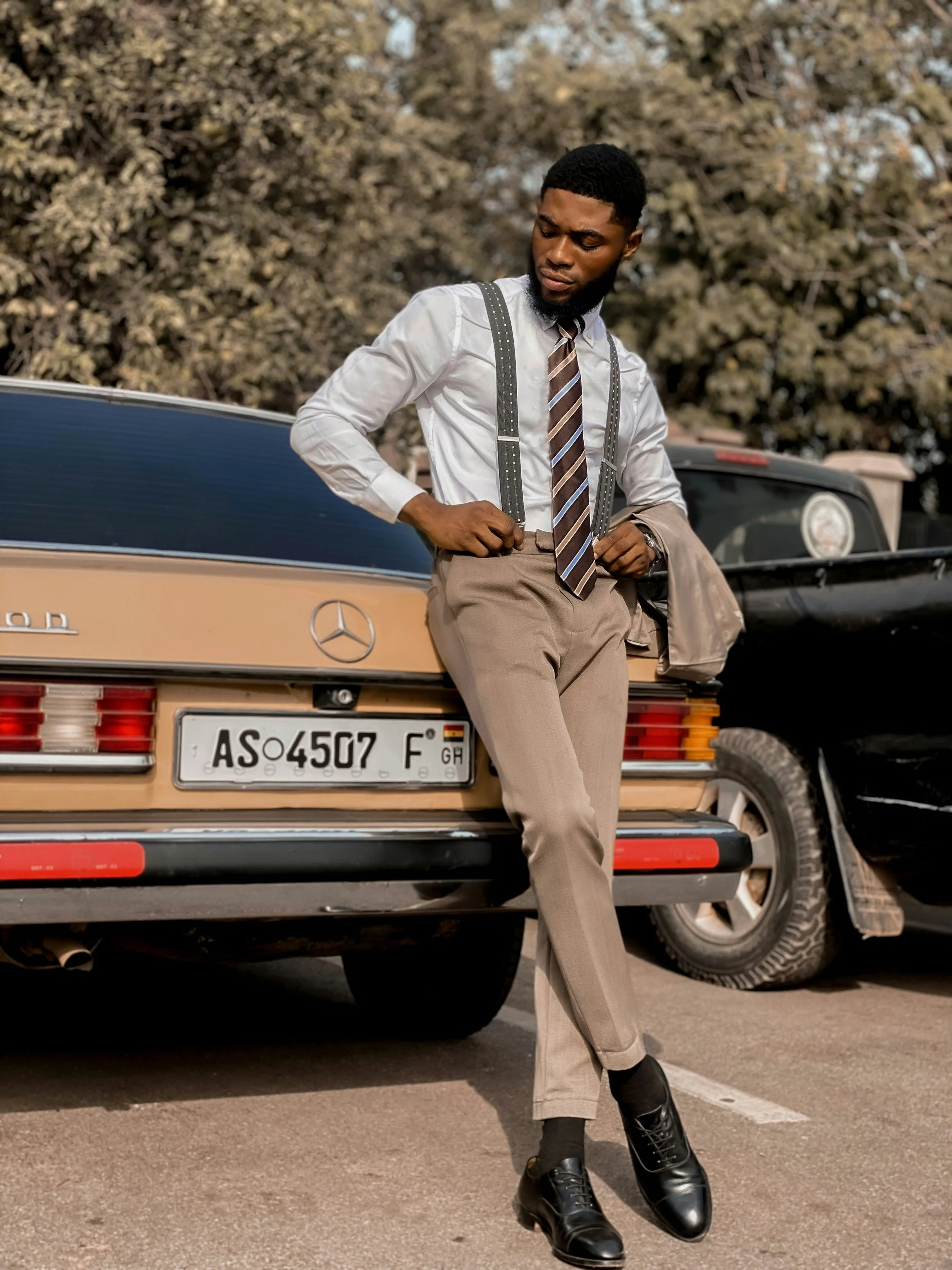 A young man in formal attire, consisting of a white shirt, striped tie, beige trousers, suspenders, and black shoes, is standing next to a vintage Mercedes-Benz car in an outdoor parking lot with trees in the background.