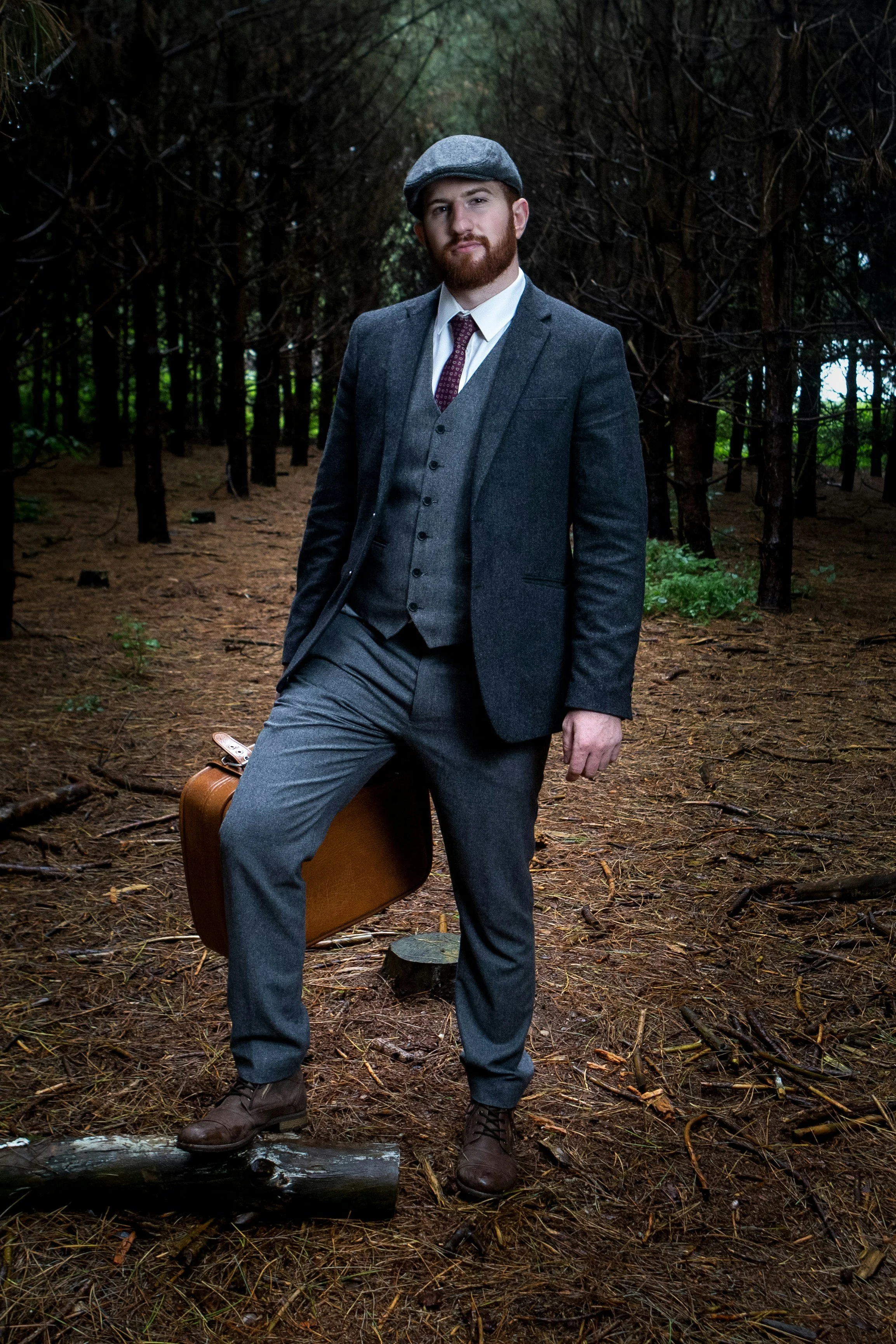 A man dressed in vintage 1920s or 1930s style suit, hat, and tie, standing on a forest trail with a suitcase, in a wooded area.