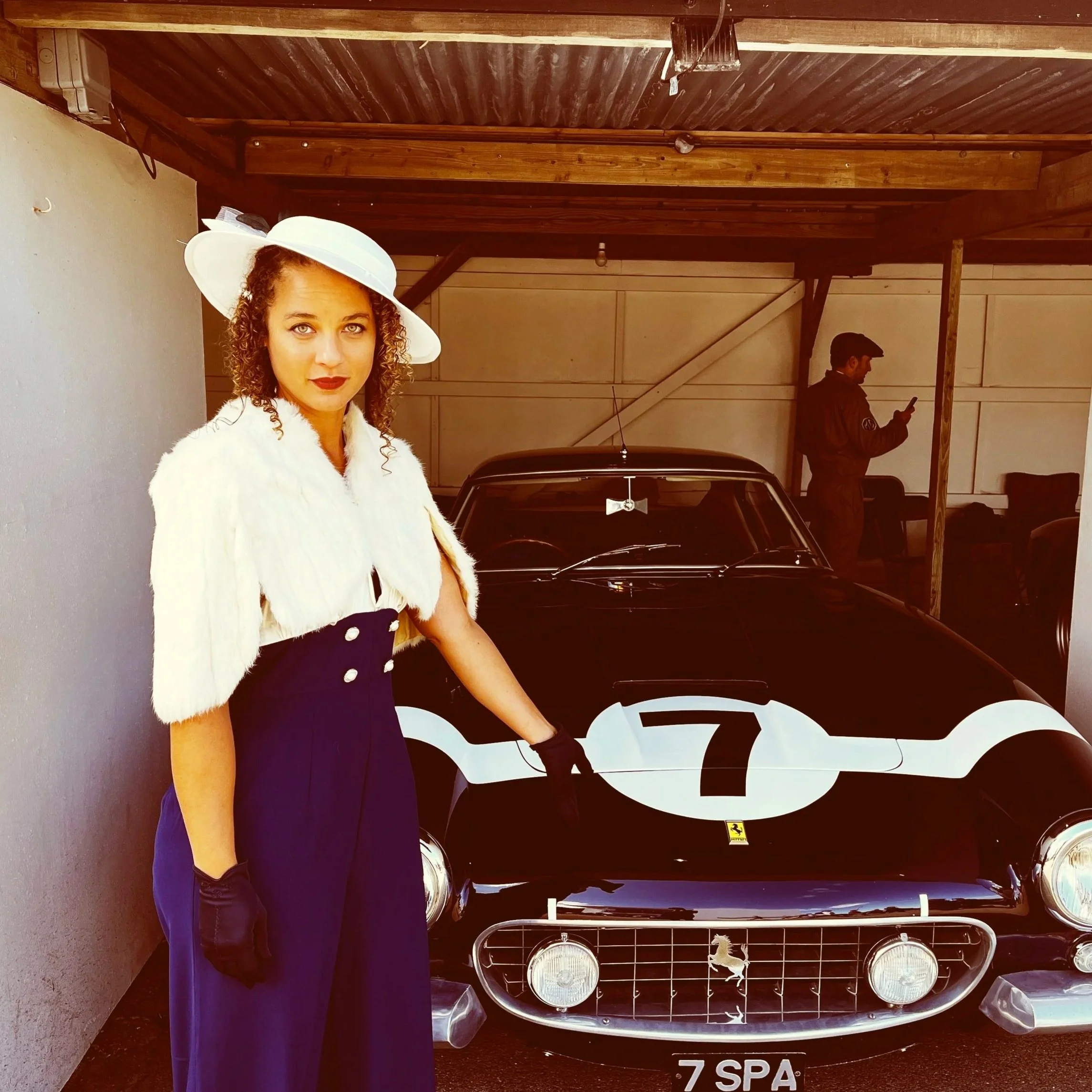 A woman dressed in vintage fashion with a white hat and fur stole stands in front of a classic black Ferrari race car, which has a large white number 7 on the hood and a Ferrari emblem on the front grille.