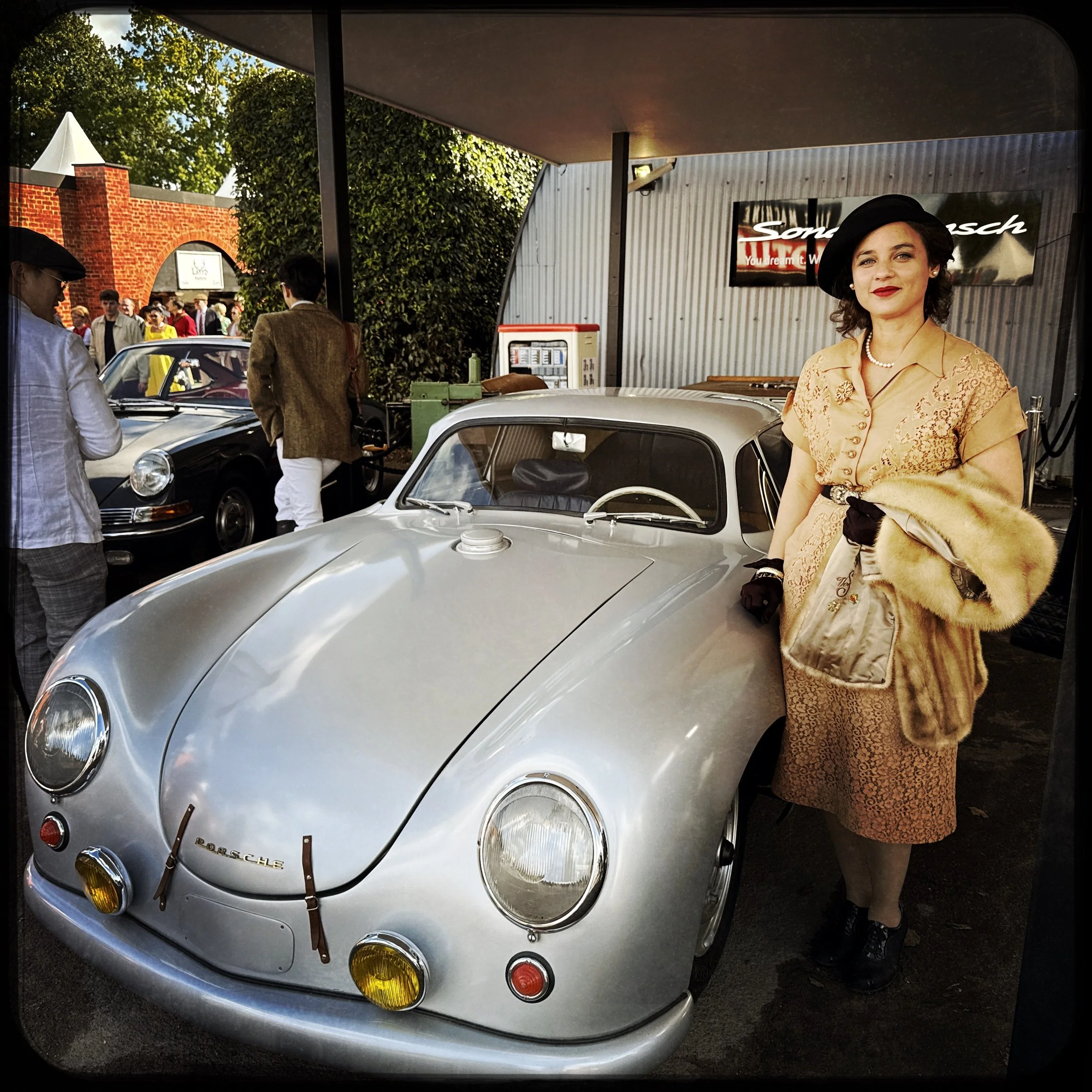 A woman dressed in vintage clothing standing next to a classic silver Porsche at a car show with other vintage cars and people in the background.