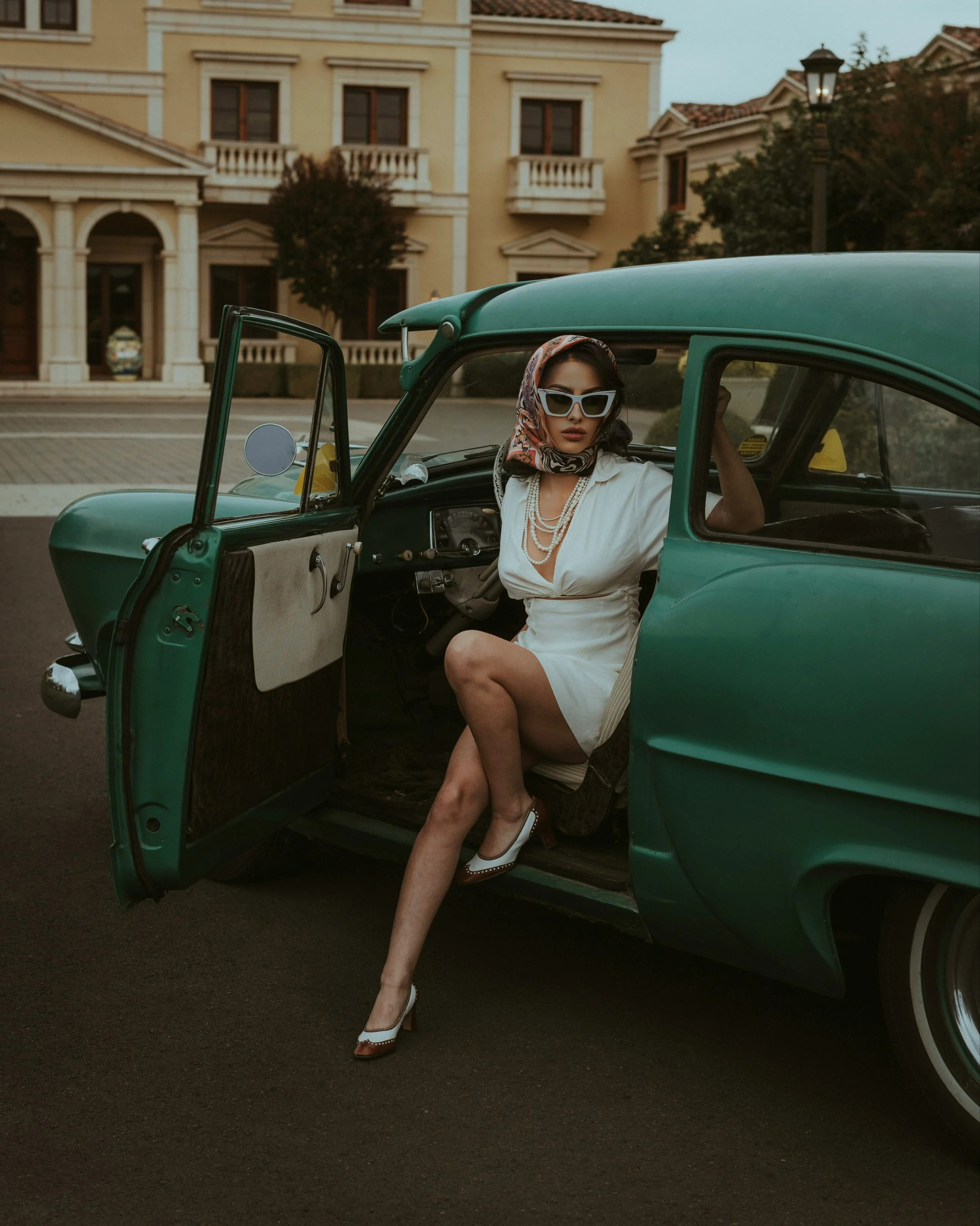A woman in a white dress, wearing sunglasses and a headscarf, sits in an open vintage green car on a city street with elegant buildings in the background.