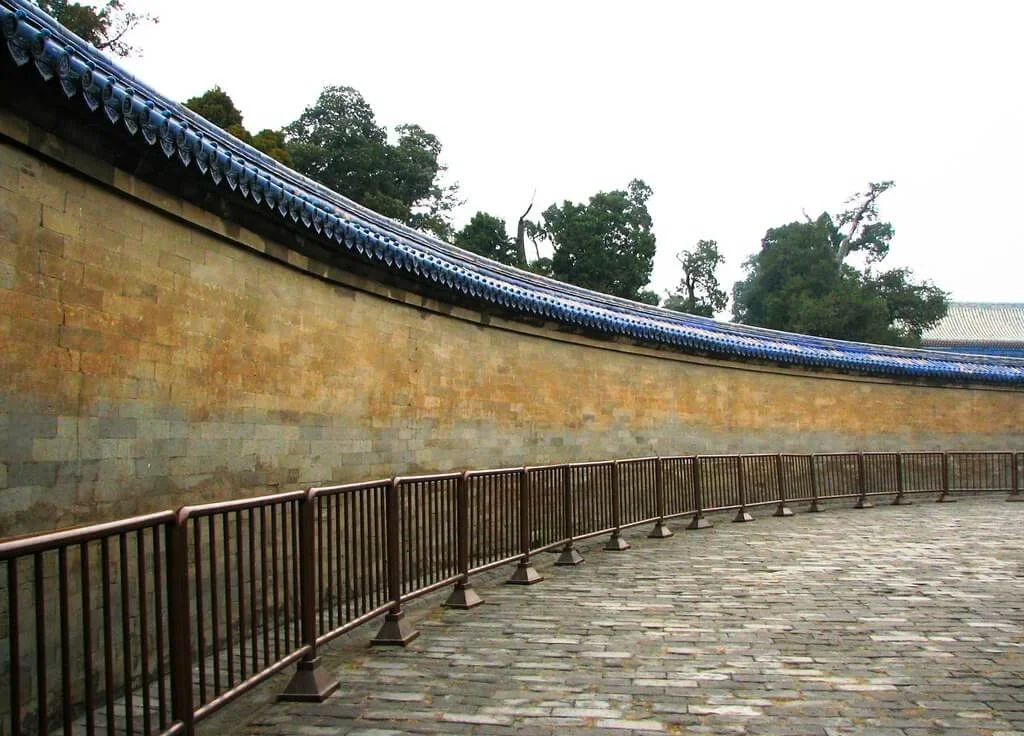 A curved stone wall with traditional Chinese blue-tiled roof, metal railing, and cobblestone pathway in a garden or historical site.