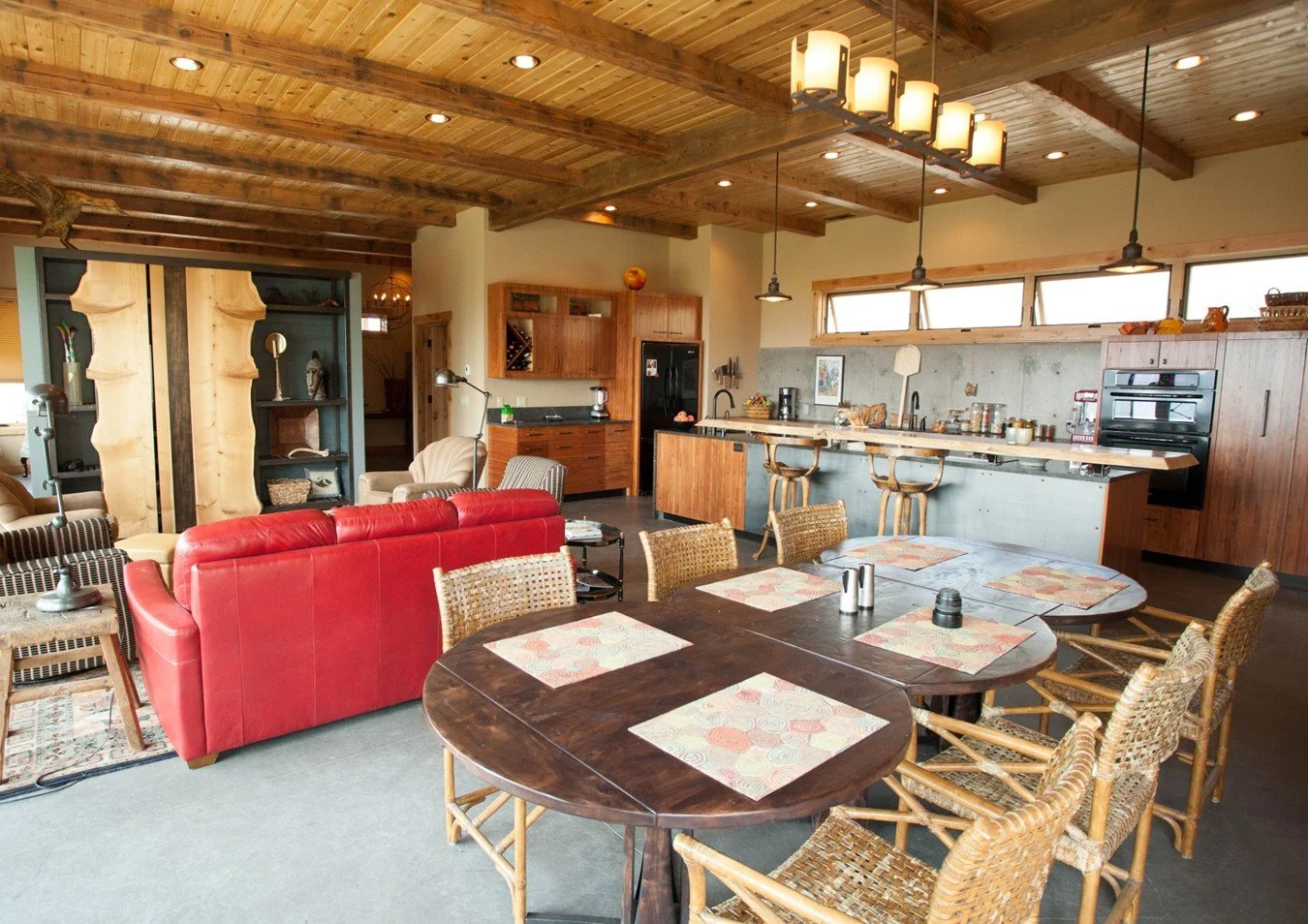 Open-concept living room and kitchen with wooden ceiling and accents, red sofa, dining table with wicker chairs, bar-style seating at kitchen island, and natural light from high windows.