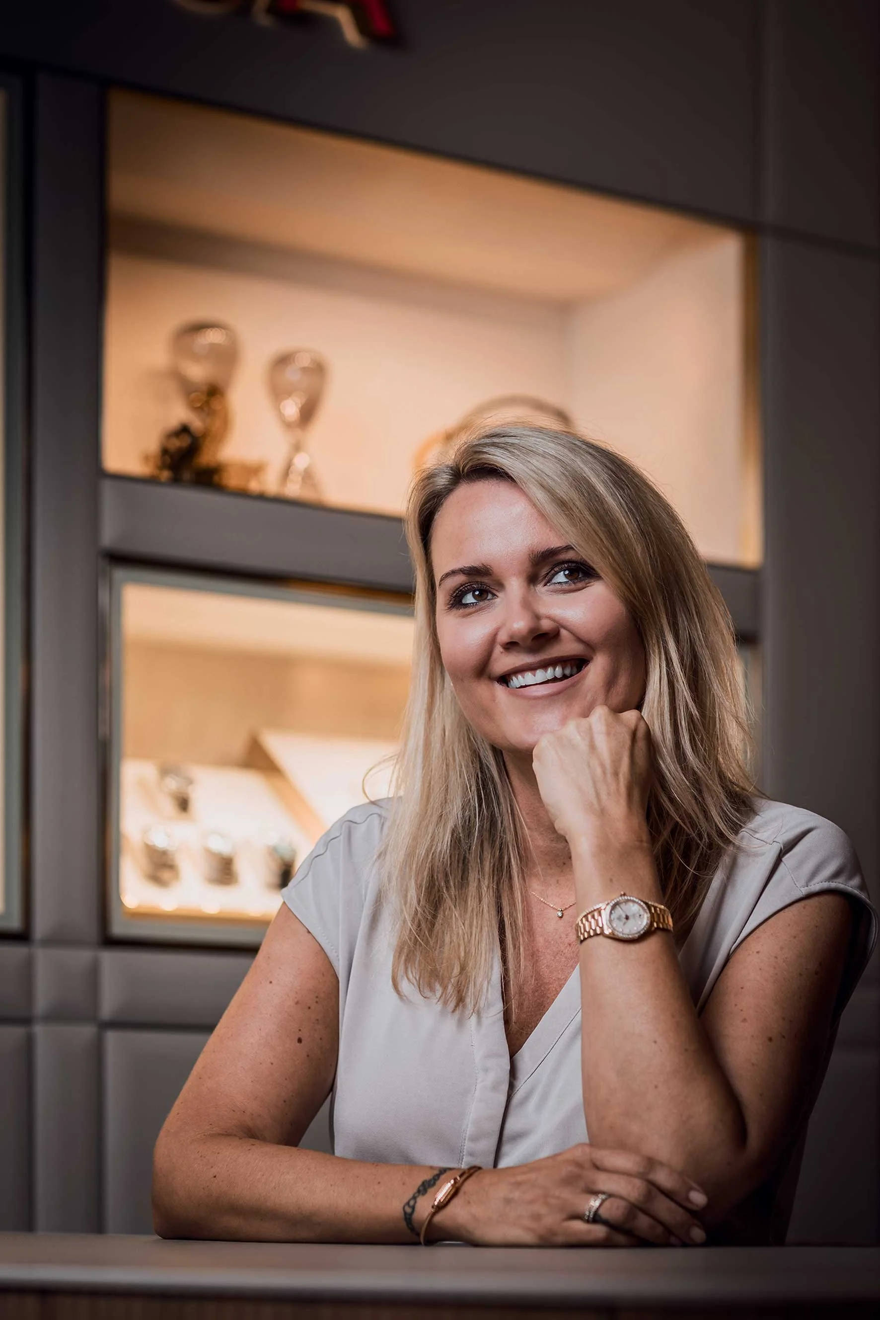 Lächelnde blonde Frau in weißer Bluse sitzt an einem Tisch und stützt ihr Kinn auf die Hand.