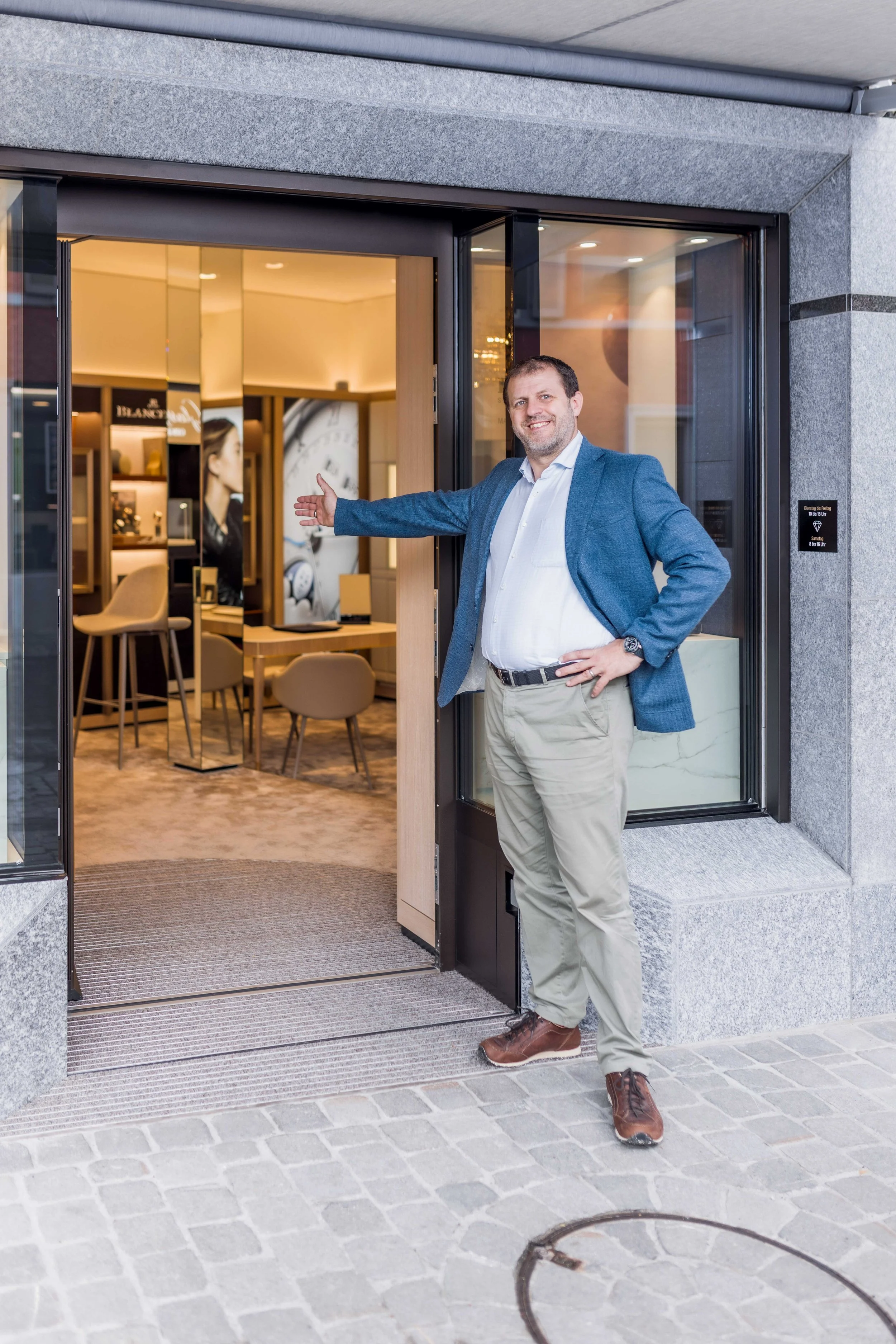 Mann im blauen Sakko präsentiert den Eingang eines eleganten Juweliergeschäfts.