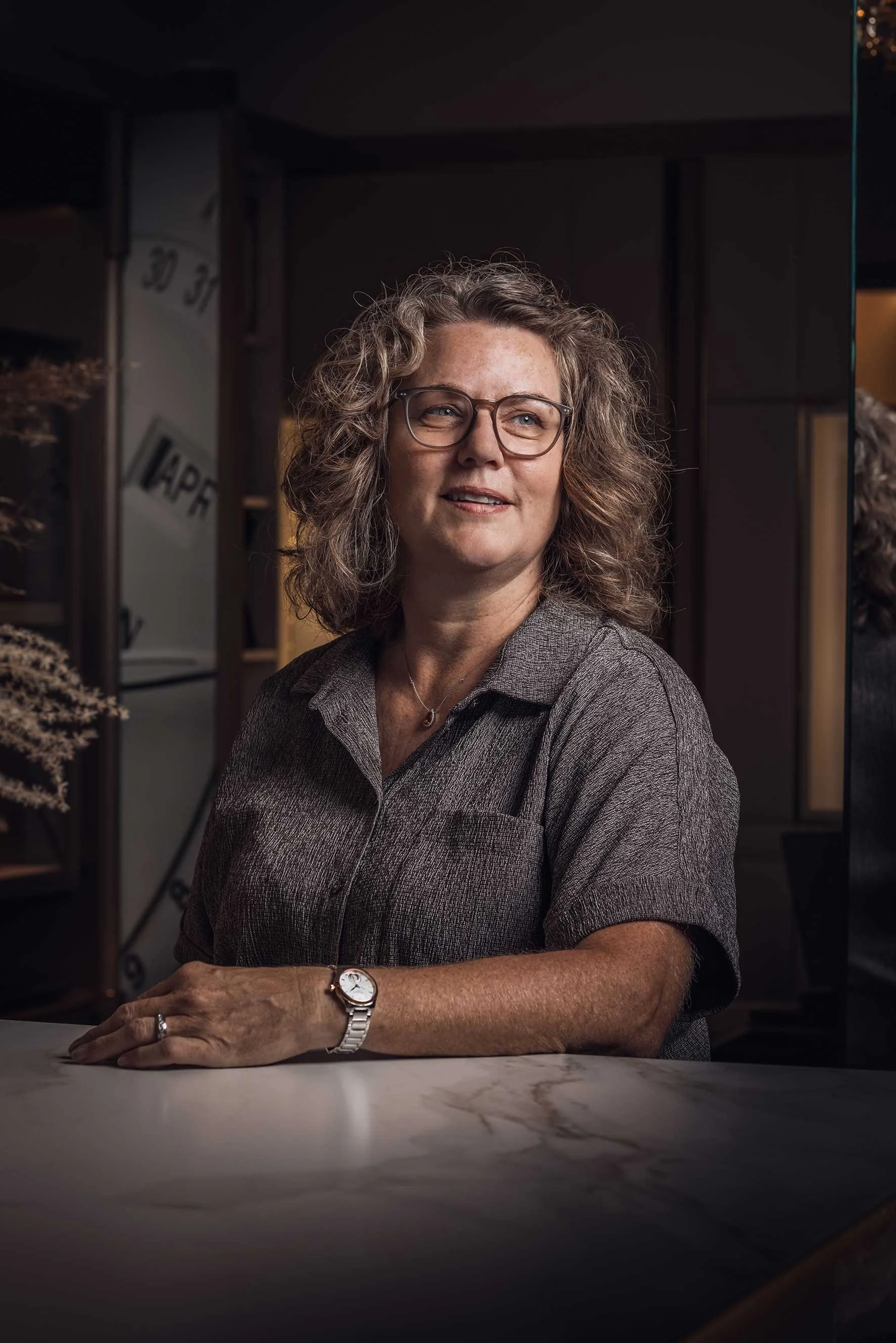 Porträt einer lächelnden Frau mit lockigem Haar und Brille, die an einem Marmortisch sitzt.