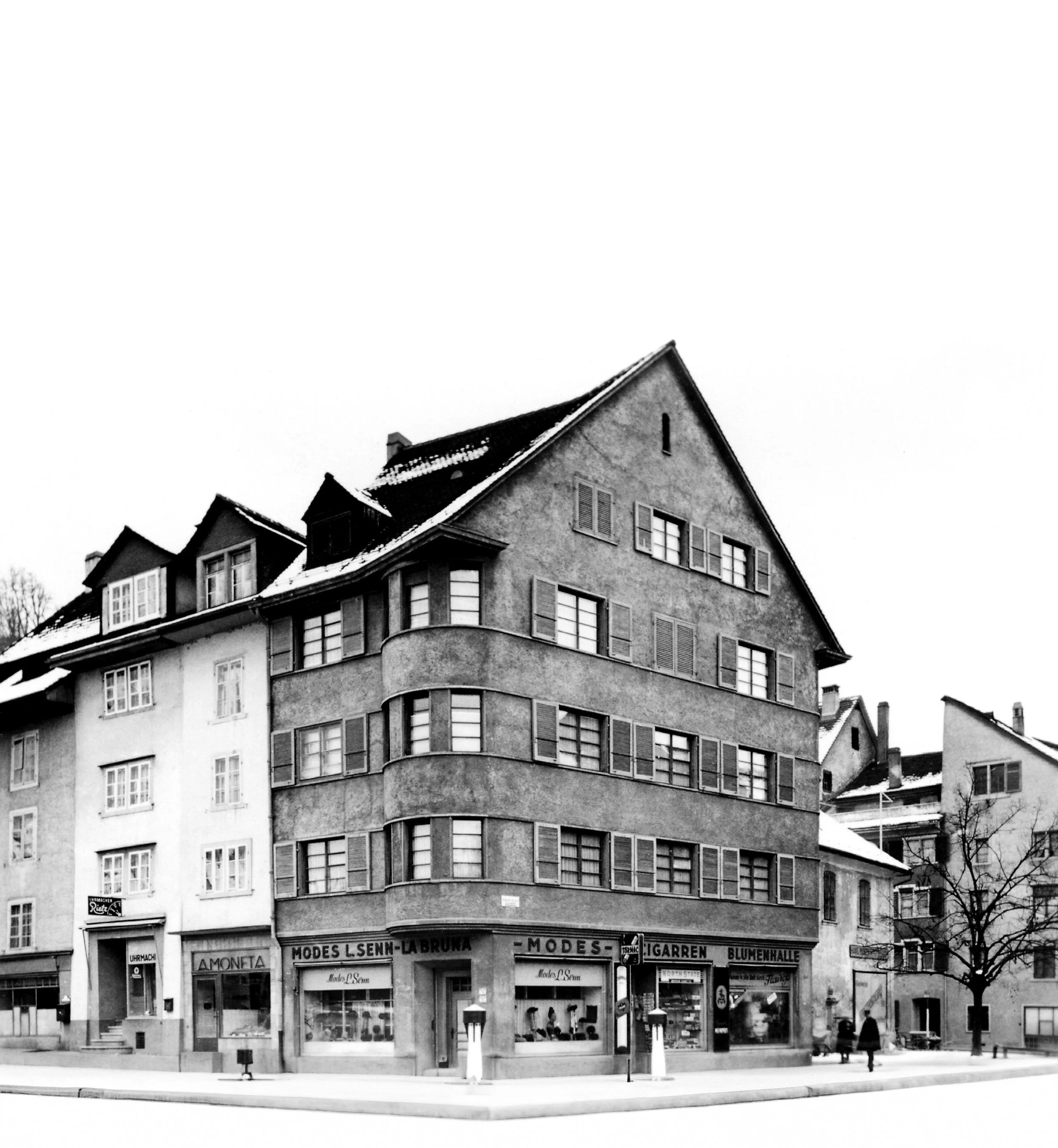 Historisches Schwarzweißfoto einer verschneiten Straßenecke mit mehrstöckigem Geschäftshaus und Ladengeschäften
