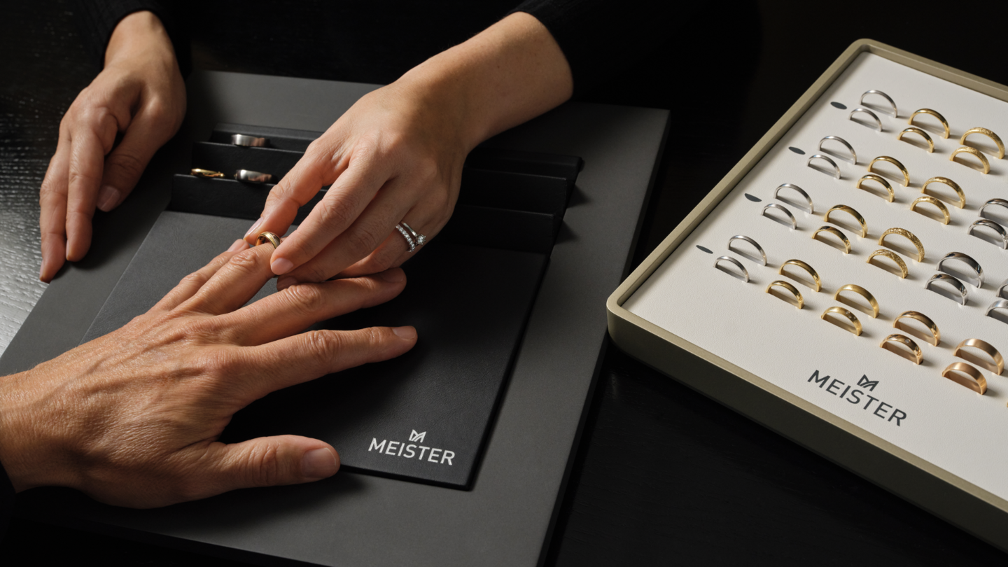 Person probiert einen goldenen Ring an; daneben präsentiert ein MEISTER-Schmuckdisplay Ringe aus Gold und Silber.