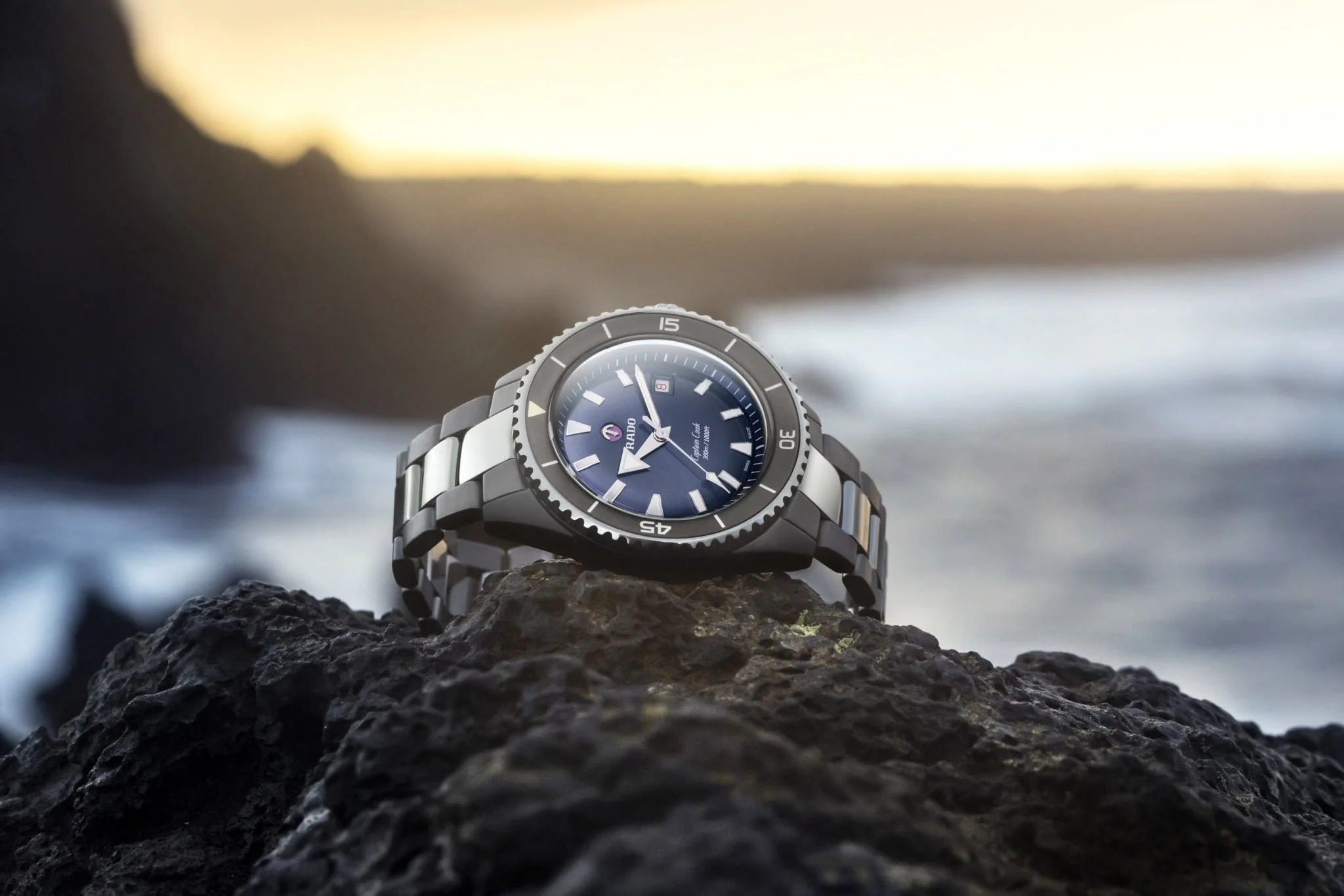 Rado Captain Cook Taucheruhr mit blauem Zifferblatt auf dunklem Felsen vor einer Küstenlandschaft bei Sonnenuntergang