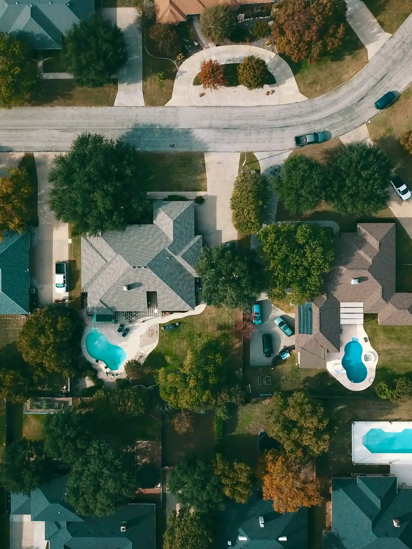 An aerial view of a residential neighborhood with houses, trees, and two swimming pools in backyards. A curving street runs through the neighborhood with parked cars.