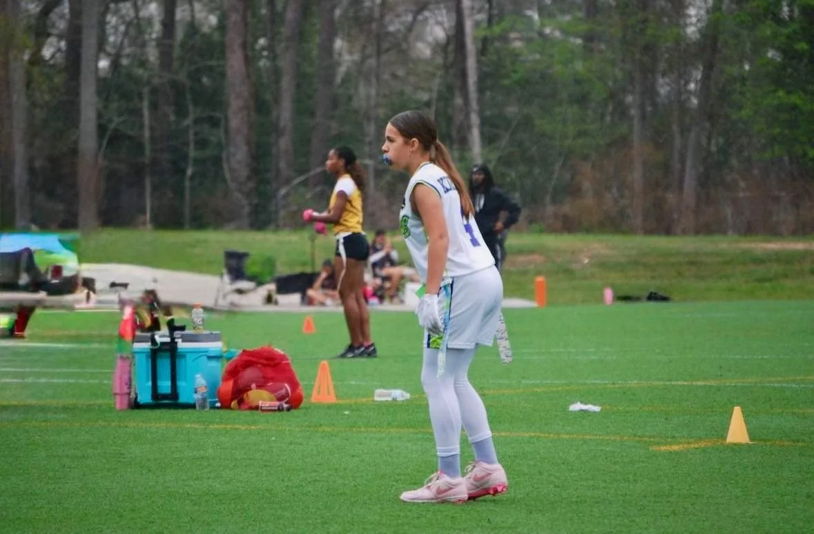 A female football player in a white uniform and pink cleats standing on the field with her hands on her hips, during practice or a game. In the background, two other women are standing, with one in a yellow shirt and black shorts and the other in a b