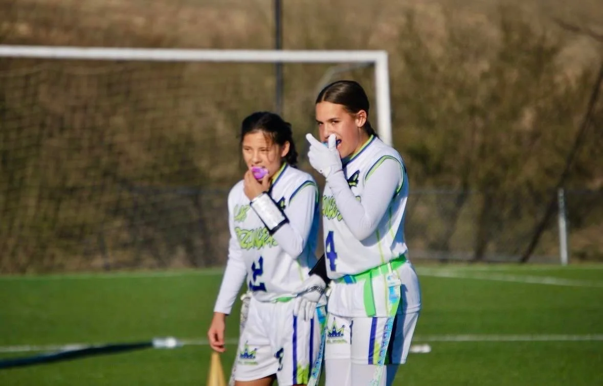 Two female flag football players standing on a field, wearing white and neon green uniforms with gloves, one with a purple mouthguard, in front of a soccer goal with a chain-link fence and trees in the background.