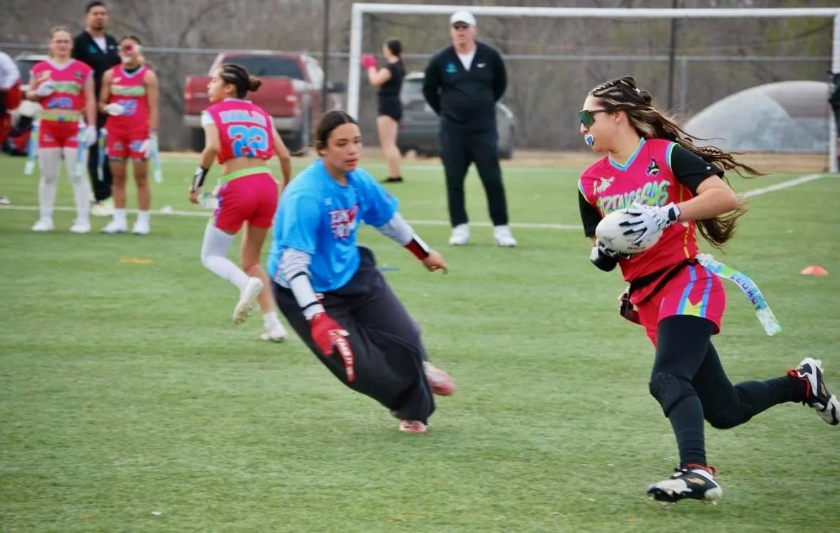 A girl in a pink and black sports uniform running with a football, while a goalie in a blue jersey attempts to stop her during a flag football game.