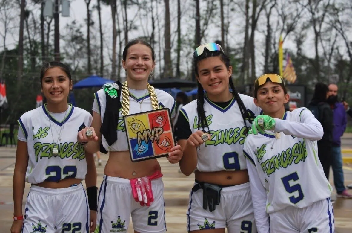 Four young girls in flag football uniforms, celebrating on an outdoor field, holding medals and a trophy, with trees and spectators in the background.