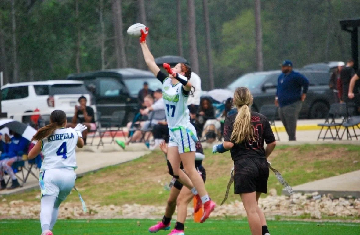 Girls playing flag football on a field with spectators and cars in the background