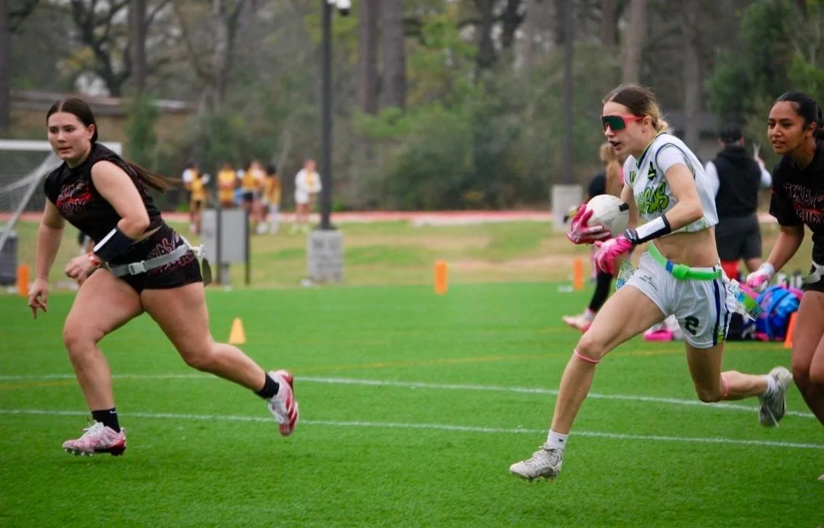 Girls playing flag football on a grassy field, one girl is running with a football while others chase her.