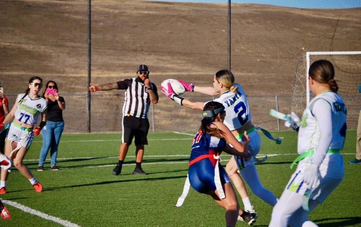 Women playing flag football, with one catching a football while being tackled by another player, and a referee with a striped shirt on a green field.