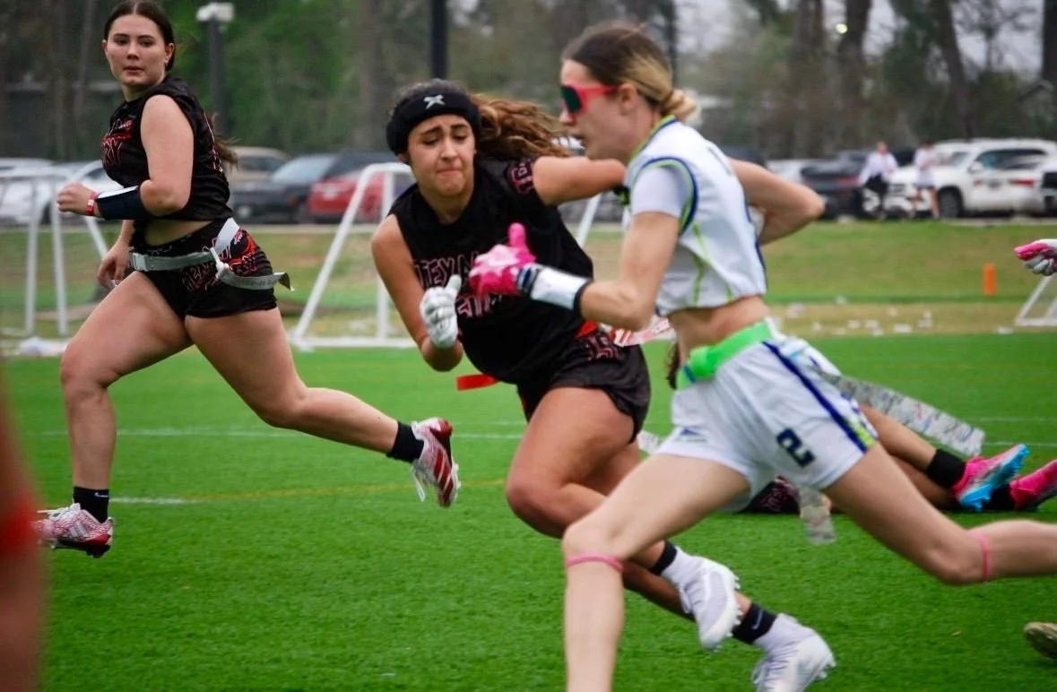 Women playing flag football on a field, with some women running and one woman in the foreground wearing pink sunglasses, a white and green uniform, and holding flags. Others are in black uniforms, chasing her.