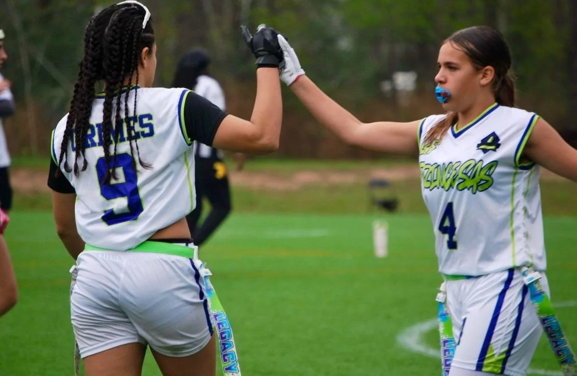 Two female flag football players wearing white and blue uniforms, high-fiving each other on a field.