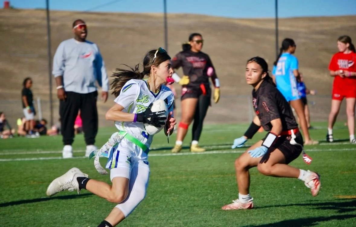 A girl in a white and green sports uniform runs with a flag football, while a girl in black sports attire positions herself on a grassy field during a flag football game, with others in the background.