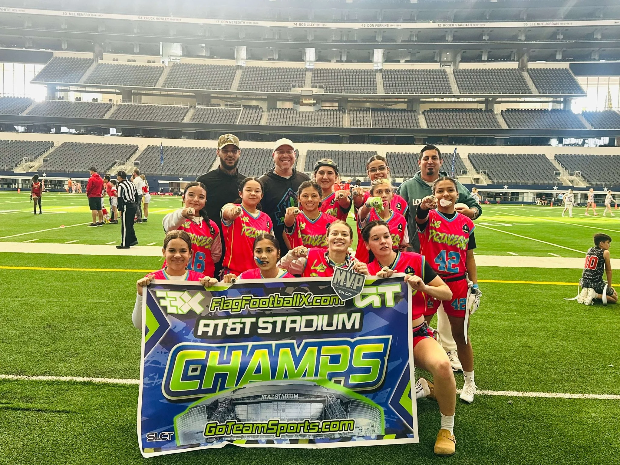 Youth girls' flag football team posing with coaches on the field holding a sign that reads 'AT&T Stadium CHAMPS' after winning a tournament, with a stadium in the background.