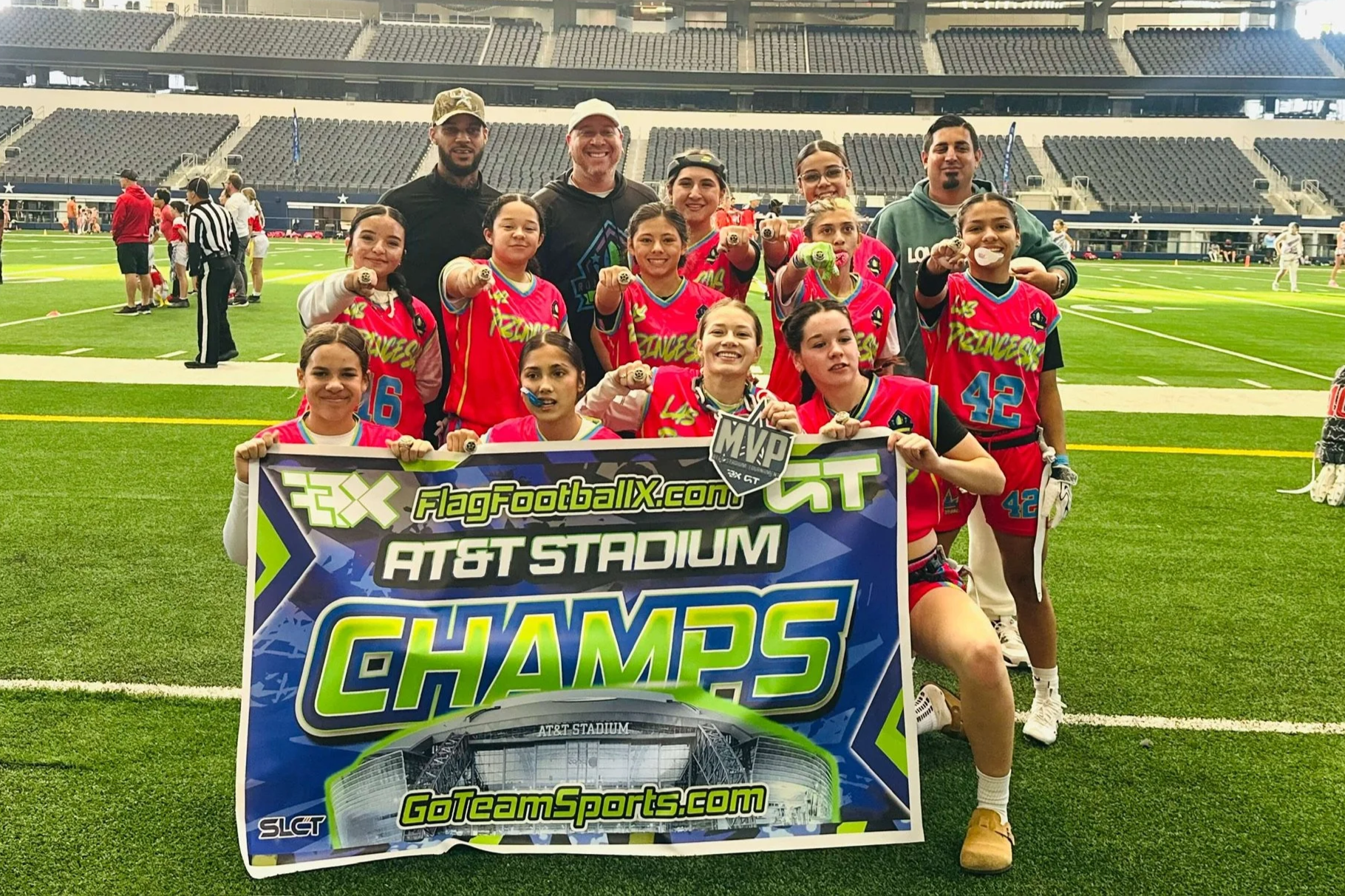 Girls' flag football team in pink uniforms celebrating on football field holding a banner that reads "FlagFootballX.com AT&T Stadium CHAMPS" with coaches behind them. Stadium seats visible in the background.