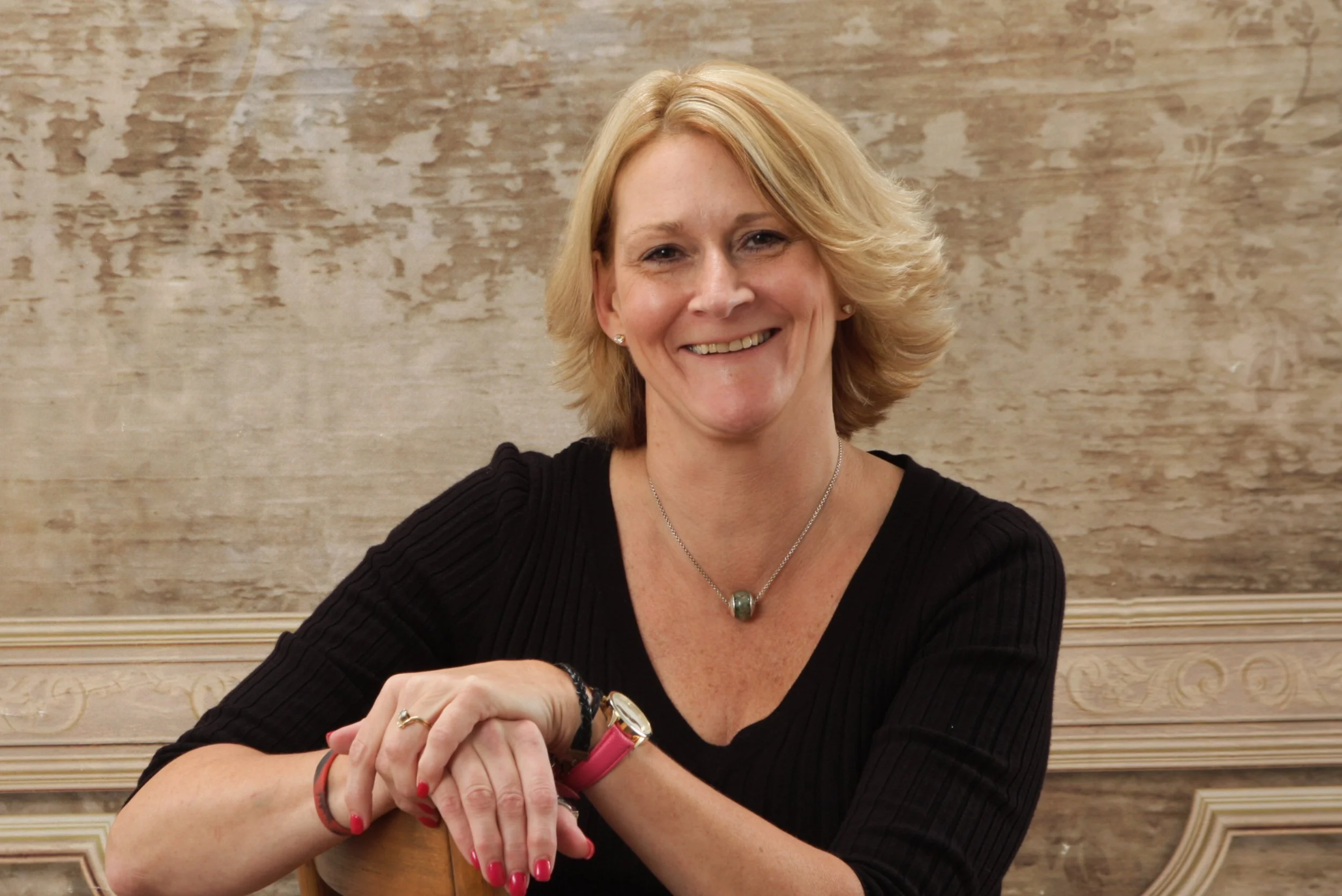 A woman with shoulder-length blonde hair, smiling, wearing a black top, jewelry, and a pink watch, sitting at a table with a textured wall background.