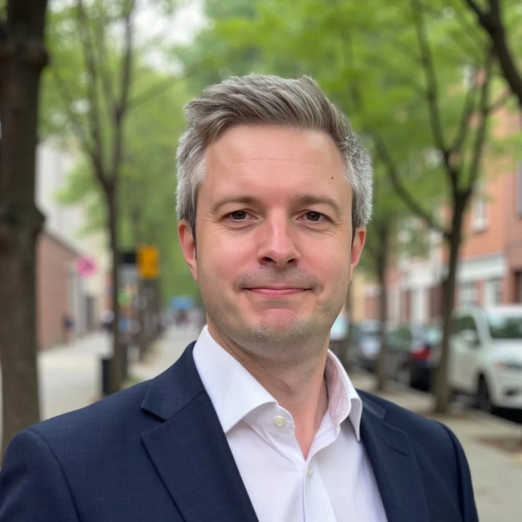 Portrait of a man with gray hair wearing a white shirt and a navy blue blazer, standing outdoors on a city street with trees and buildings in the background.
