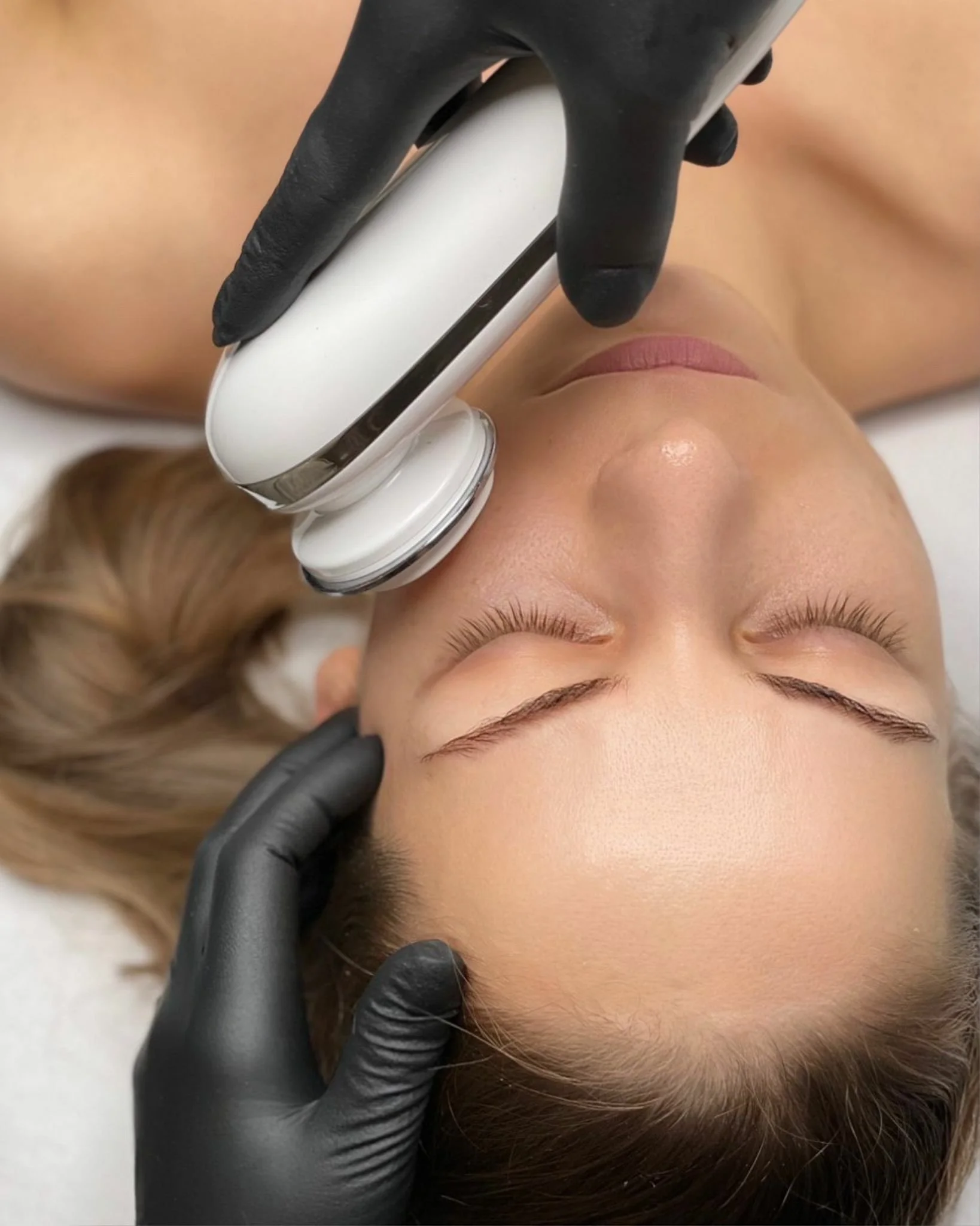 A person receiving a facial treatment using a white handheld device, lying on a treatment bed, with closed eyes, while wearing black gloves.