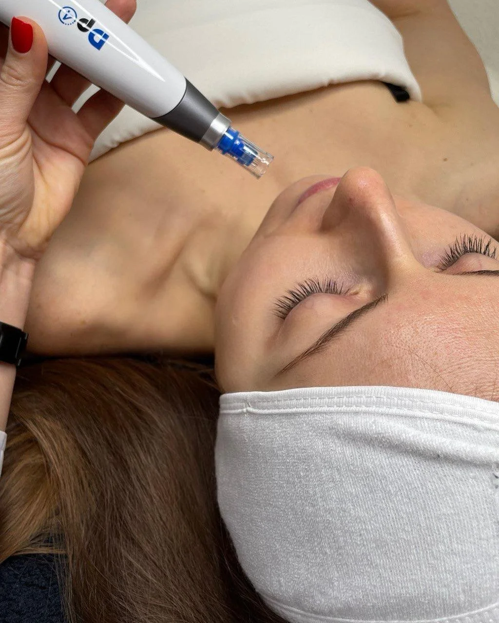 A woman receiving a cosmetic or skincare treatment with a handheld device on her face, lying down with a white head covering.
