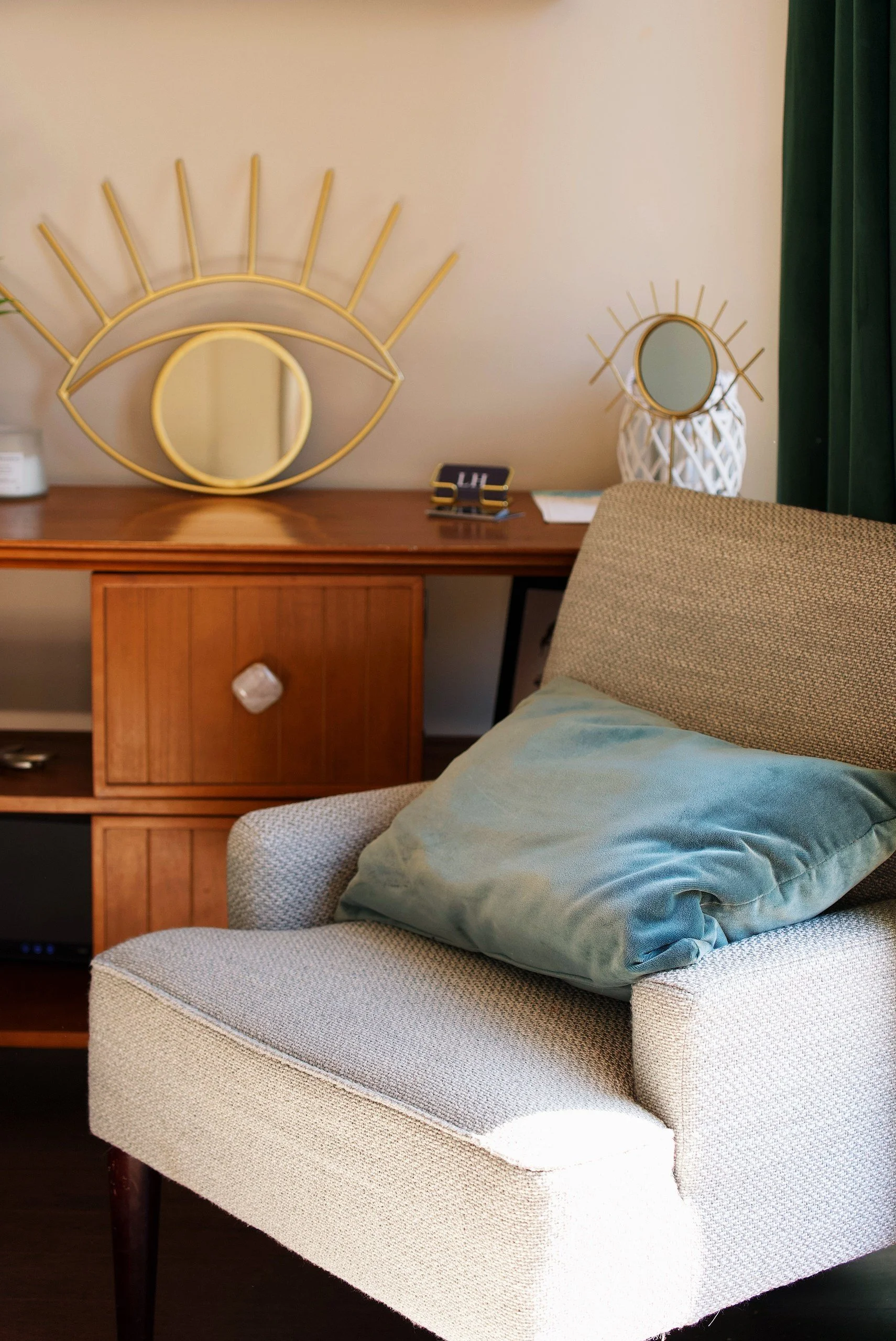 A beige upholstered armchair with a light blue pillow resting on its seat. Behind it, a wooden side table with a white yarn lamp, a small framed photo, and a paper. Two decorative mirrors are on the wall above the table, one shaped like an eye with a circular mirror for the iris, the other round with radiating lines. A dark green curtain is on the right side.
