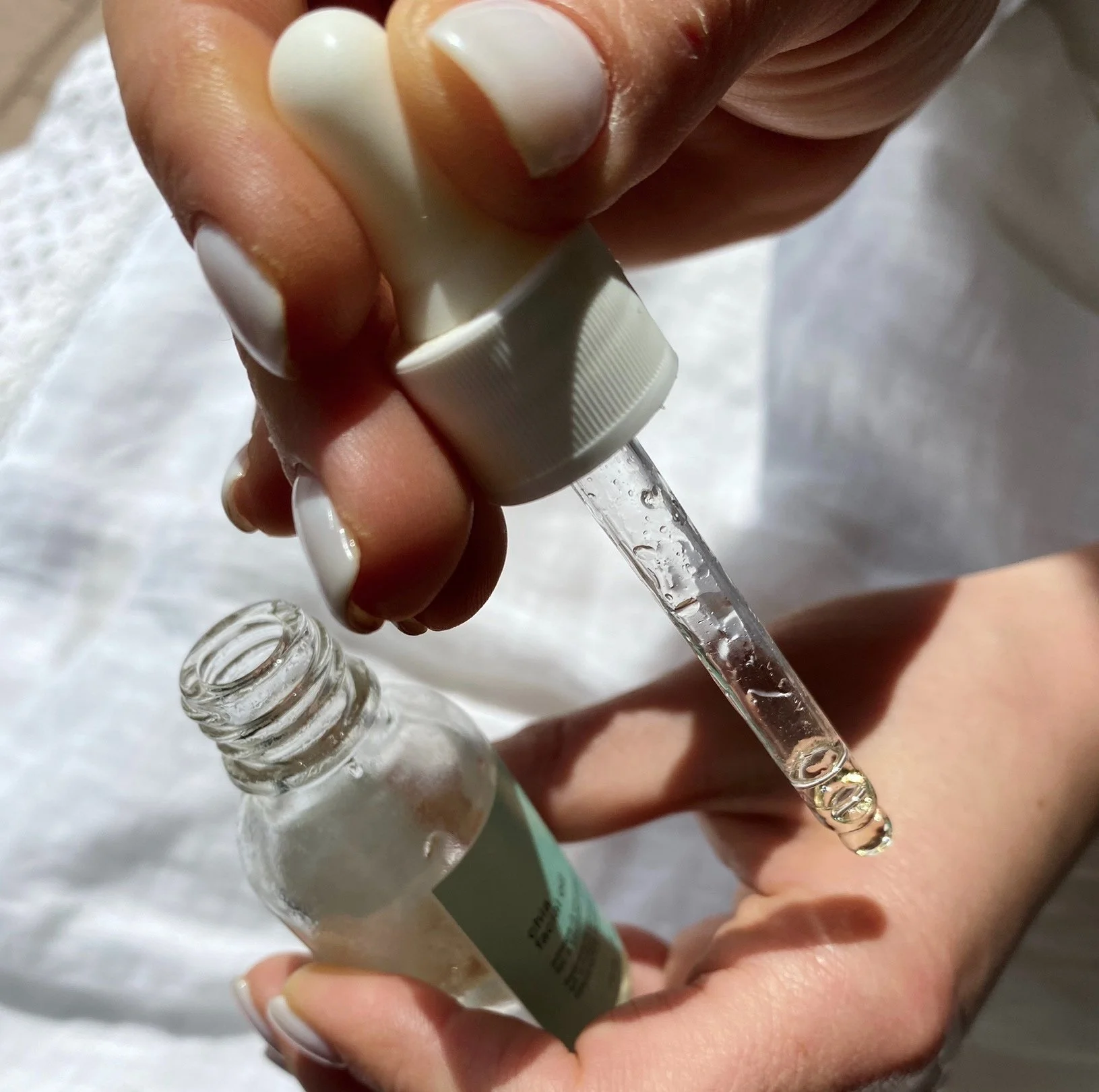 A person is holding a glass dropper and a small bottle, transferring a clear liquid into the dropper, with the bottle's open mouth visible. The person has well-manicured nails with white nail polish, and the background appears to be a white or light-colored surface.