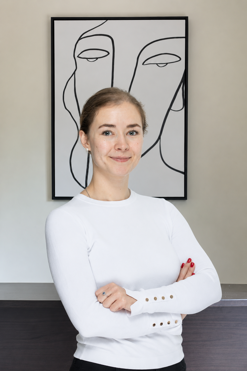 A woman with light brown hair, blue eyes, wearing a white long-sleeve shirt with buttons on the sleeves, standing with arms crossed in front of a black and white line art portrait of a face on a wall.