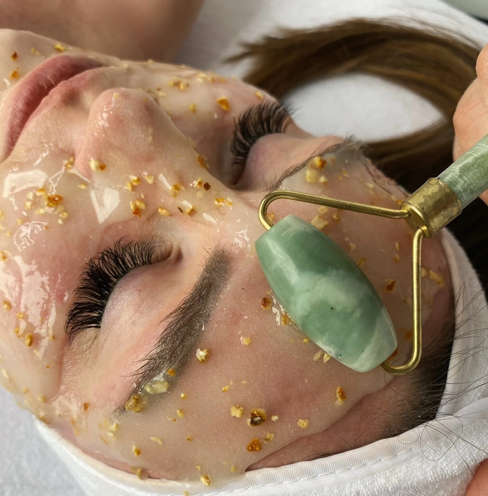 Close-up of a woman receiving a facial treatment with a jade roller, her face covered in a thick, yellowish facial mask and small particles.