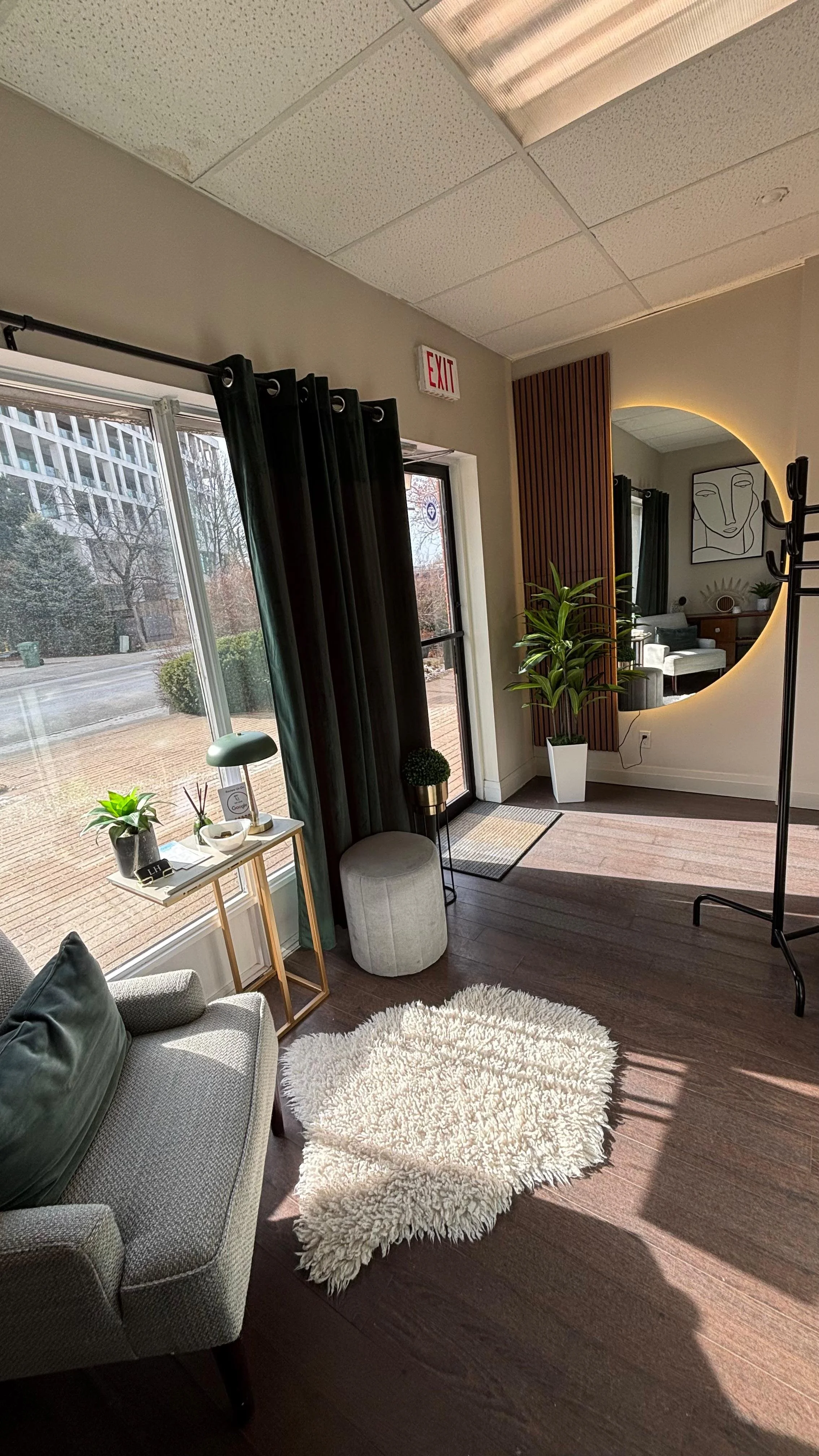 A cozy living room corner with a light grey armchair, dark green cushions, and a fluffy white rug on wood flooring. Beside the armchair is a small white side table with a potted plant, a lamp, and decorative items. Large windows with dark curtains allow natural light, and a large round mirror with a backlight hangs on the wall. There are indoor plants and modern artwork, creating a stylish and inviting space.