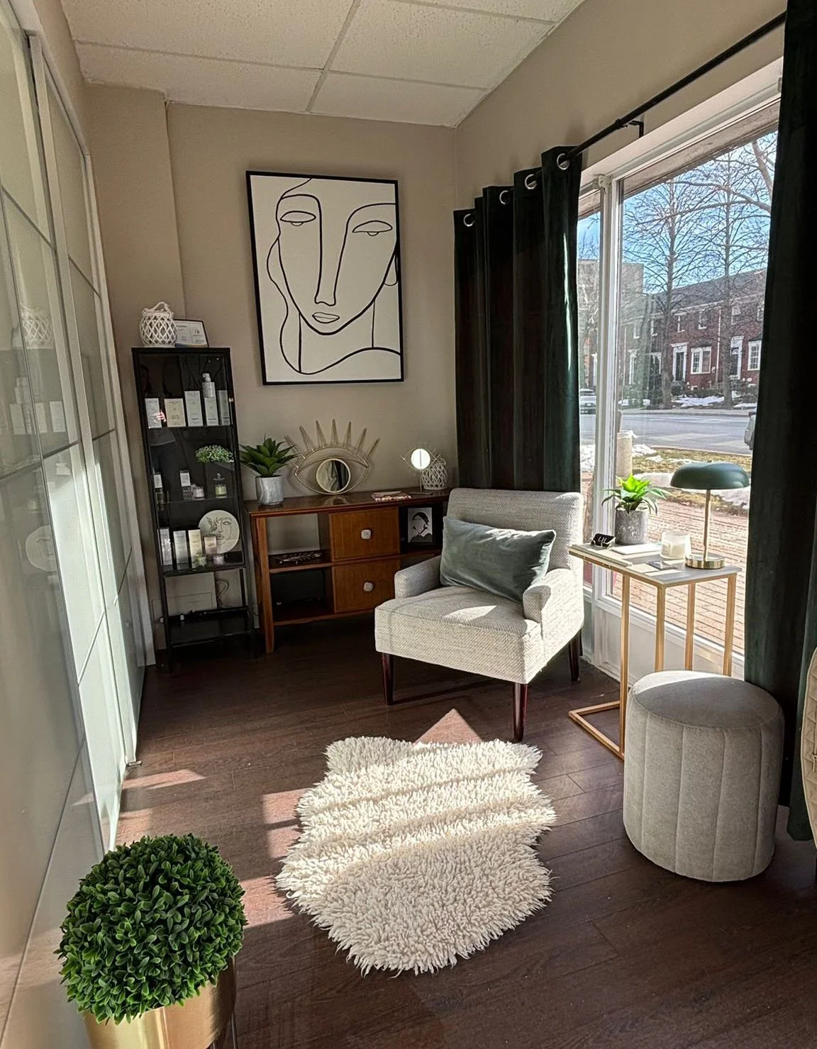 A cozy living room corner with a white armchair, a small side table with a green plant and a decorative lamp, a black and glass shelving unit with various decor and skincare products, a large abstract line art portrait on the wall, a wooden sideboard with a plant and decor items, a white fluffy rug, and a window with black curtains showing a street view with bare trees and brick houses.