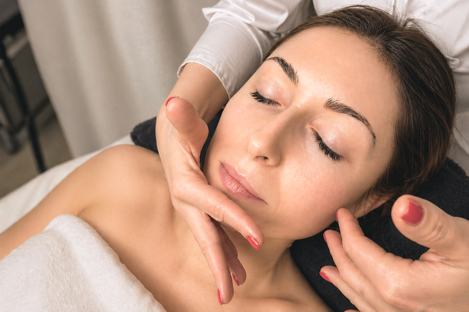 A woman receiving a facial massage in a spa setting, lying on a treatment bed with her eyes closed.