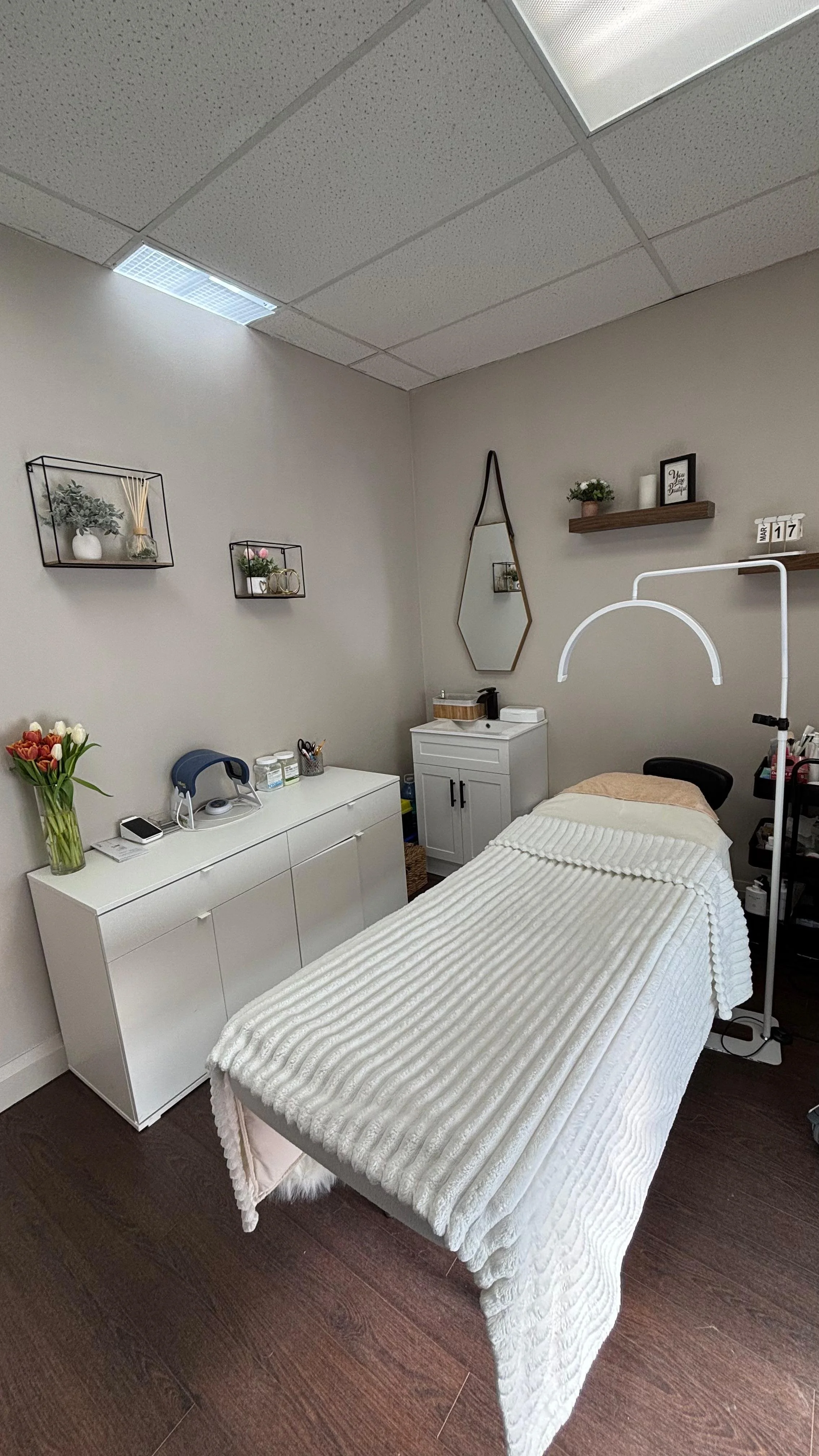A medical or wellness treatment room with a massage or examination bed covered in a cream-colored, ribbed blanket. There is a white cabinet with a vase of flowers, a phone, and a blue device on top. The wall has decorative shelves with plants, a mirror, and framed messages. The ceiling has fluorescent and acoustic tiles, and the room has dark hardwood flooring.