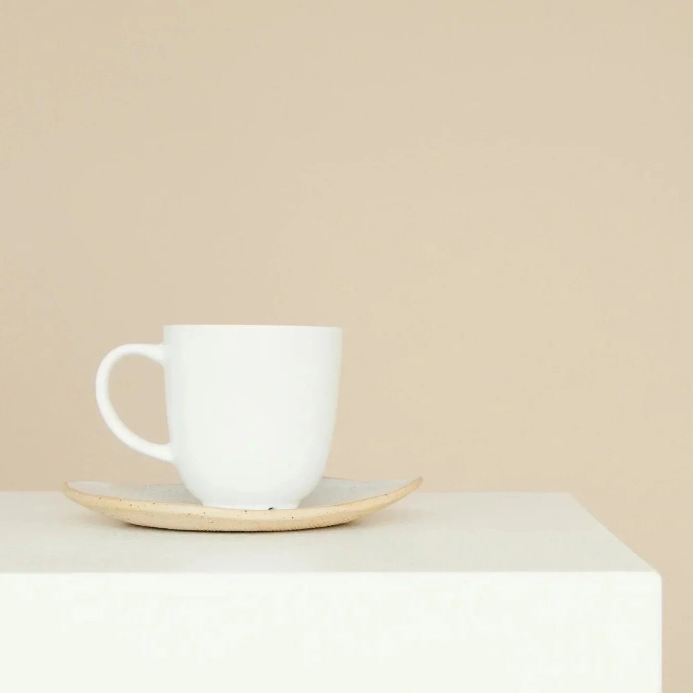 White ceramic mug on a beige saucer, on a white surface against a beige background.