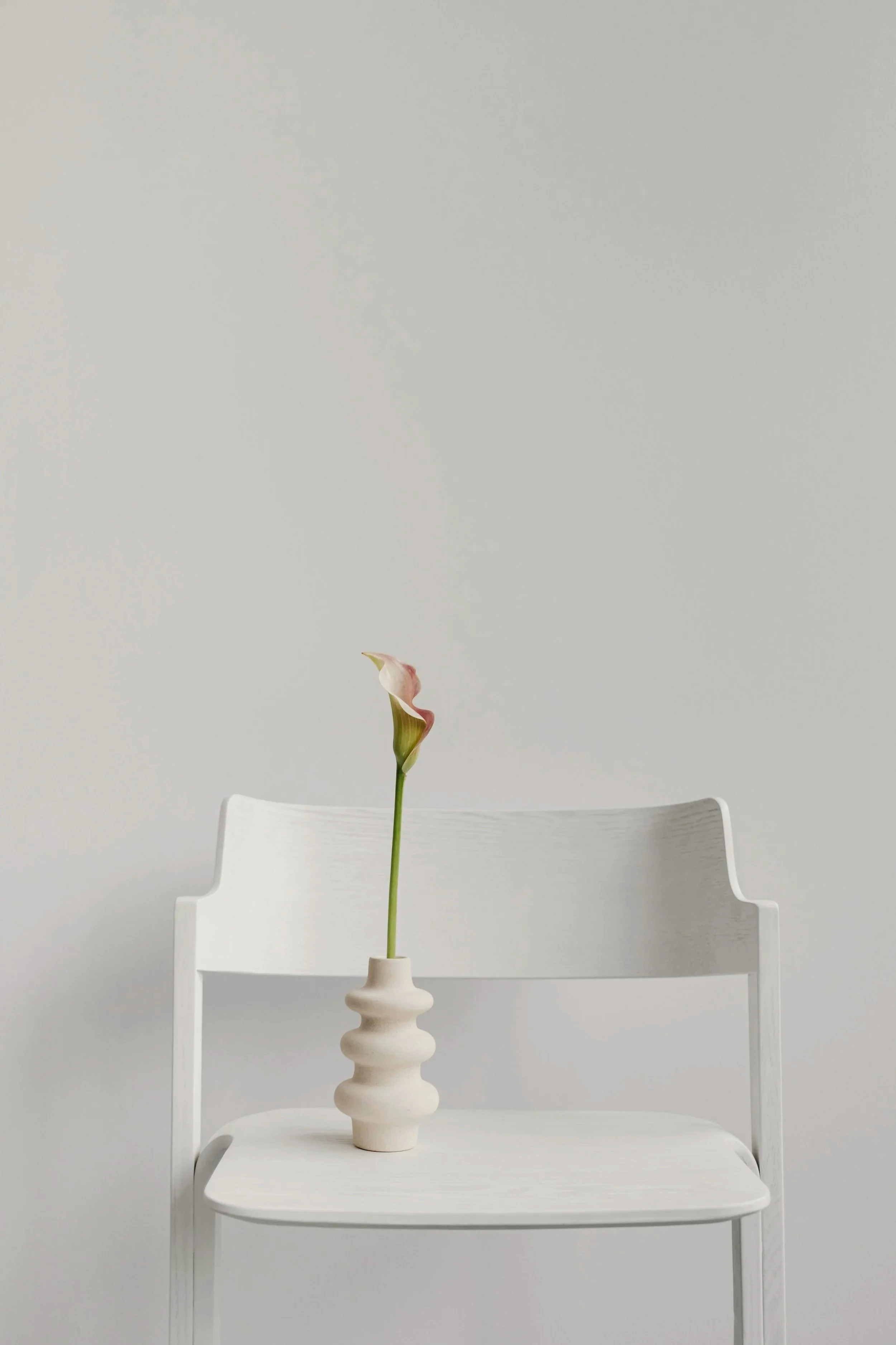 A white chair with a tall backrest, positioned in front of a plain light gray wall. On the chair's seat sits a white, wavy ceramic vase containing a single tall flower with a long green stem and pink, cream, and white petals.