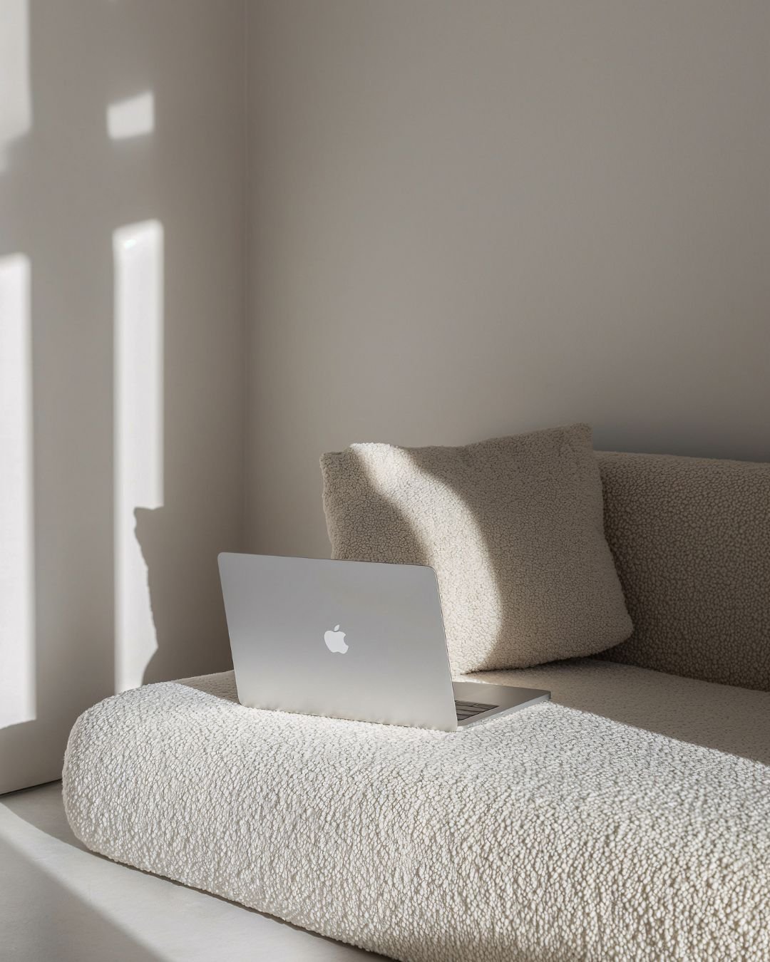 A silver MacBook on a textured beige sofa with a matching beige pillow, sunlight creating shadow patterns on the wall and sofa.