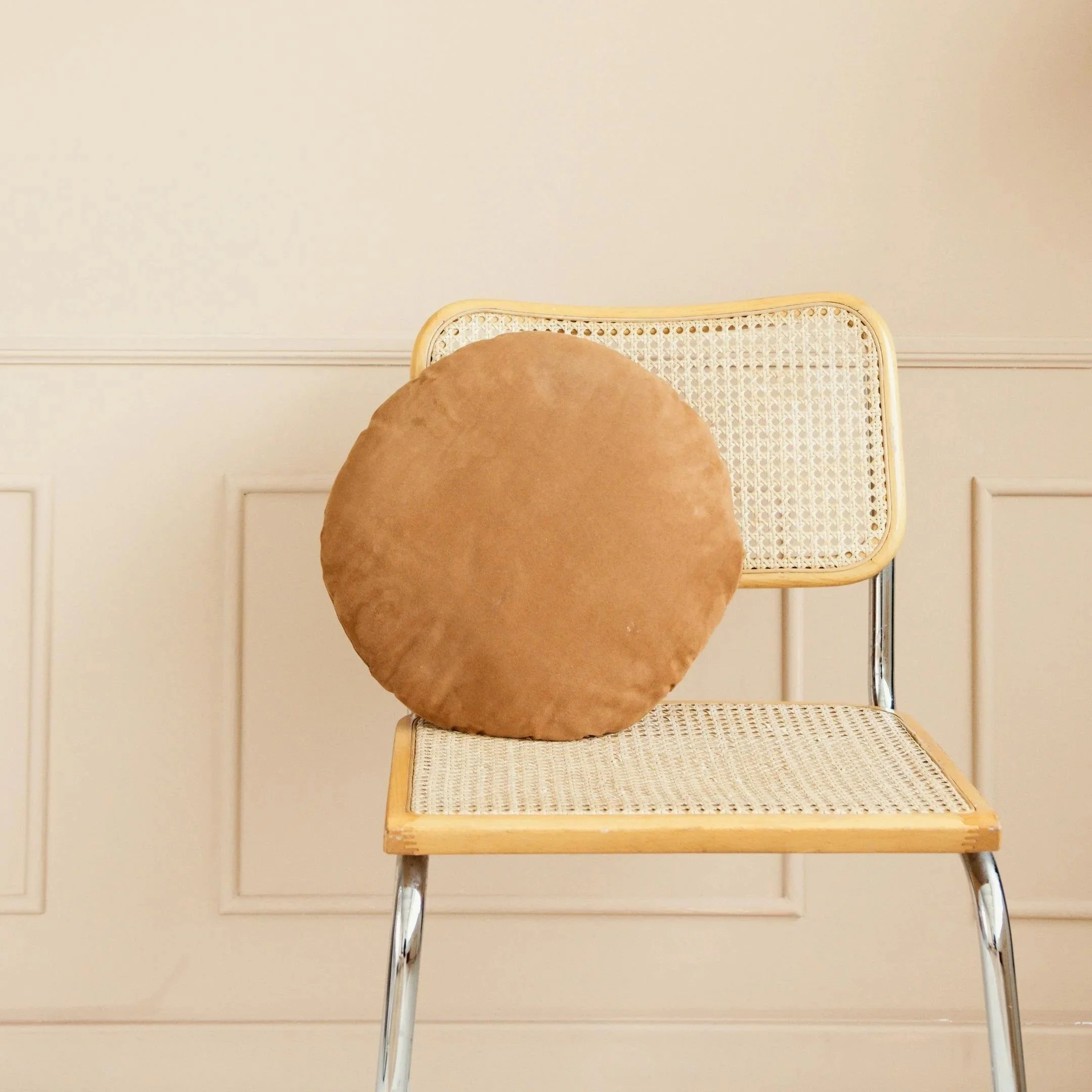 A beige cushioned chair with a woven cane backrest and metal legs positioned in front of a white wall with decorative molding.