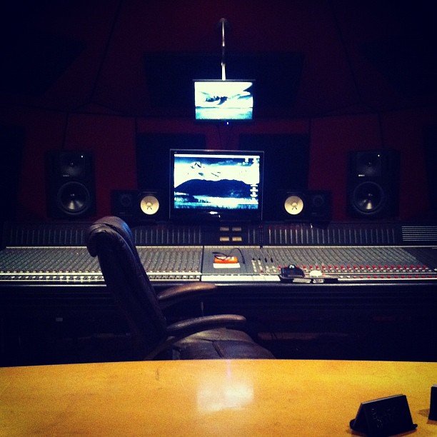 A professional recording studio with a mixing console, large speakers, and computer screens displaying scenic images, with a black leather chair in front.