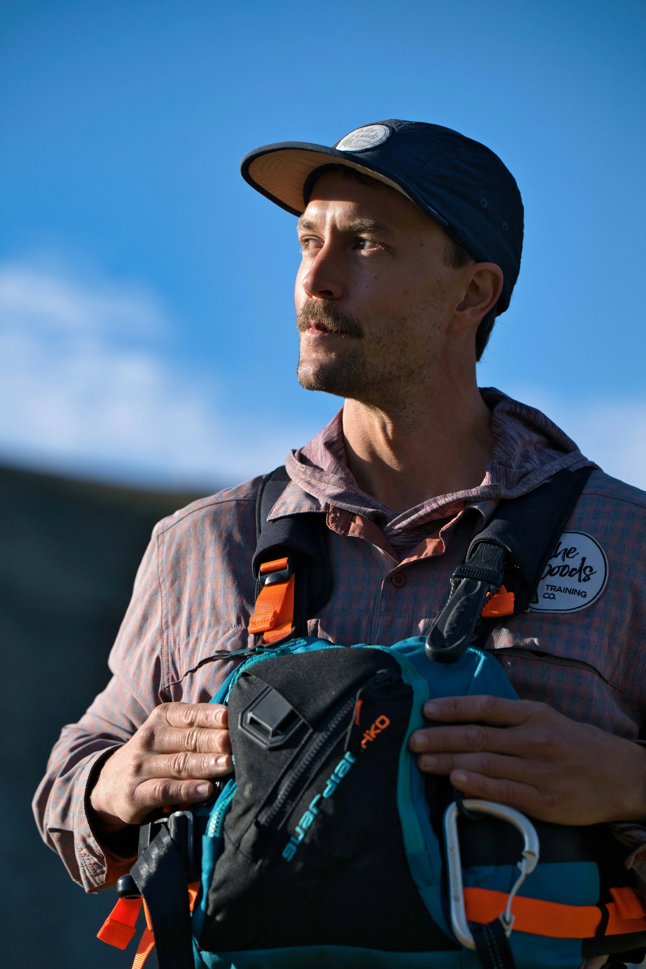 A man wearing a blue cap, plaid shirt, and a PFD with orange straps, outdoors with a clear blue sky.